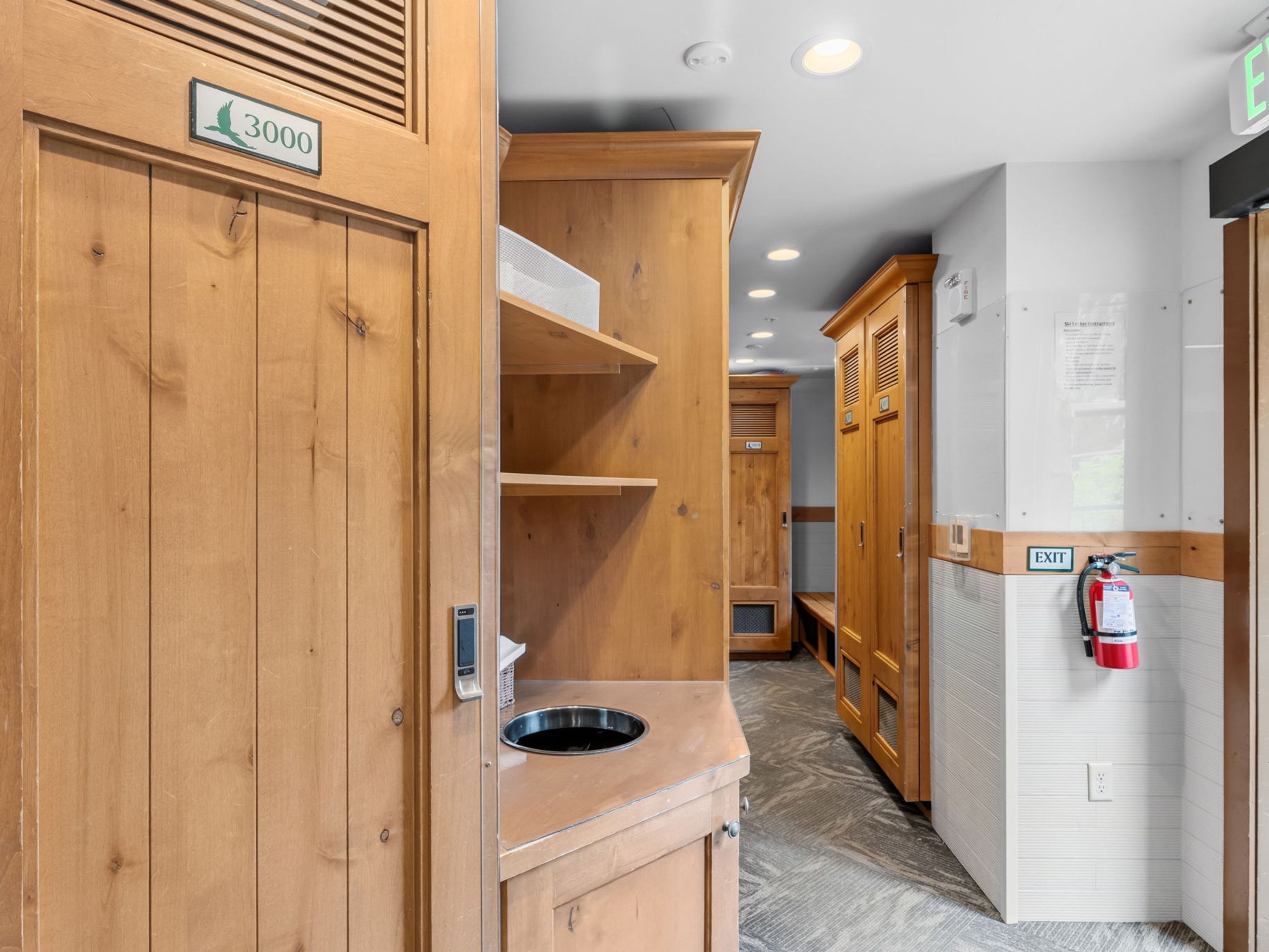 This image captures a locker room or ski storage area featuring light wood cabinetry and lockers with louvered details. A small, built-in sink area with open shelving sits in the foreground, while a fire extinguisher is mounted on the wall near an exit sign. The space has a functional, utilitarian design with patterned flooring and recessed ceiling lighting.