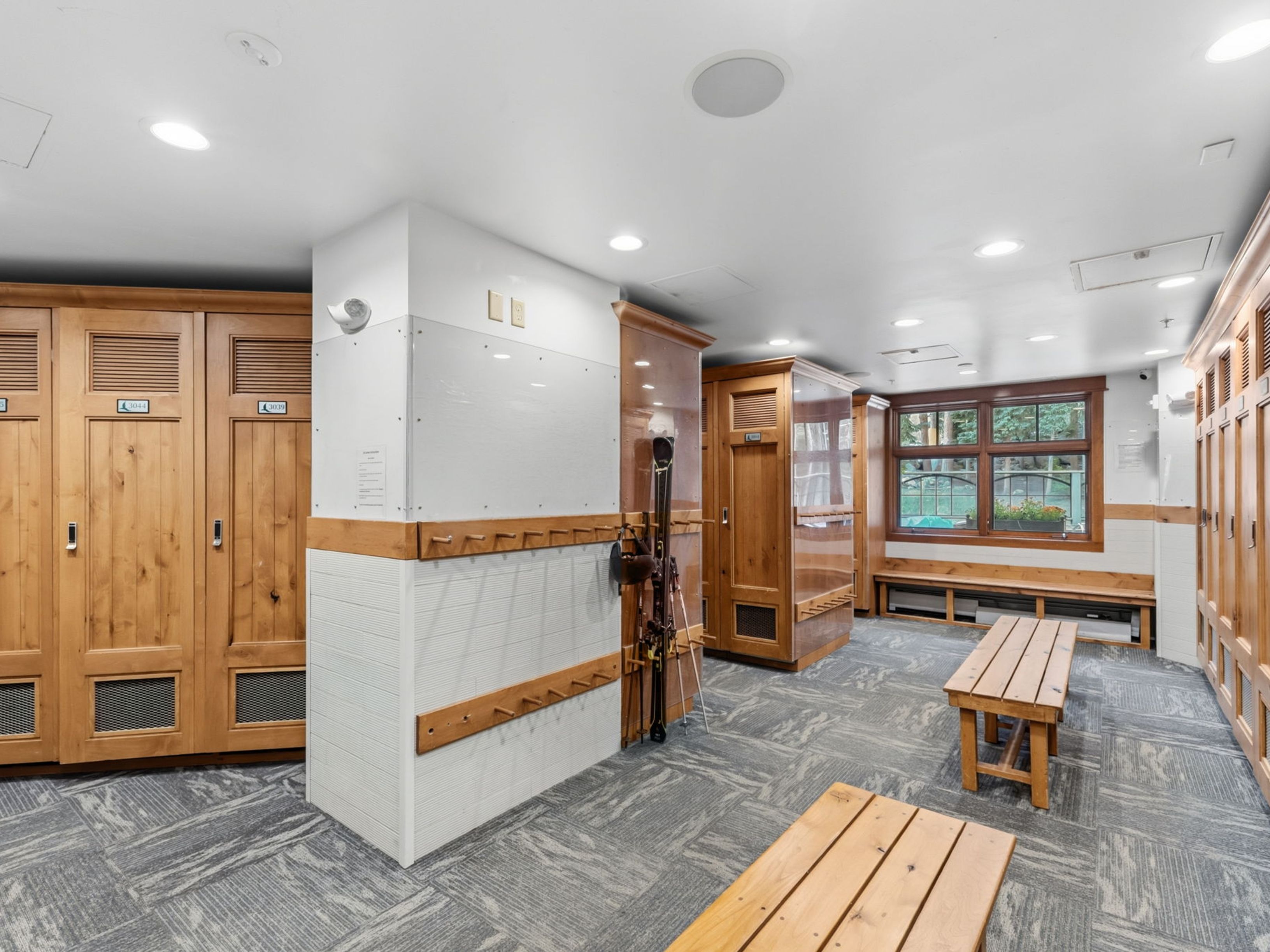 This image depicts a ski locker room, likely located in a resort or residential complex, featuring rows of light wood lockers with integrated ventilation grilles. The space is equipped with wooden benches for changing, a wall-mounted rack for skis, and a neutral patterned carpet. The room is brightly lit by recessed ceiling lights and includes a window that provides a view of the outdoors.