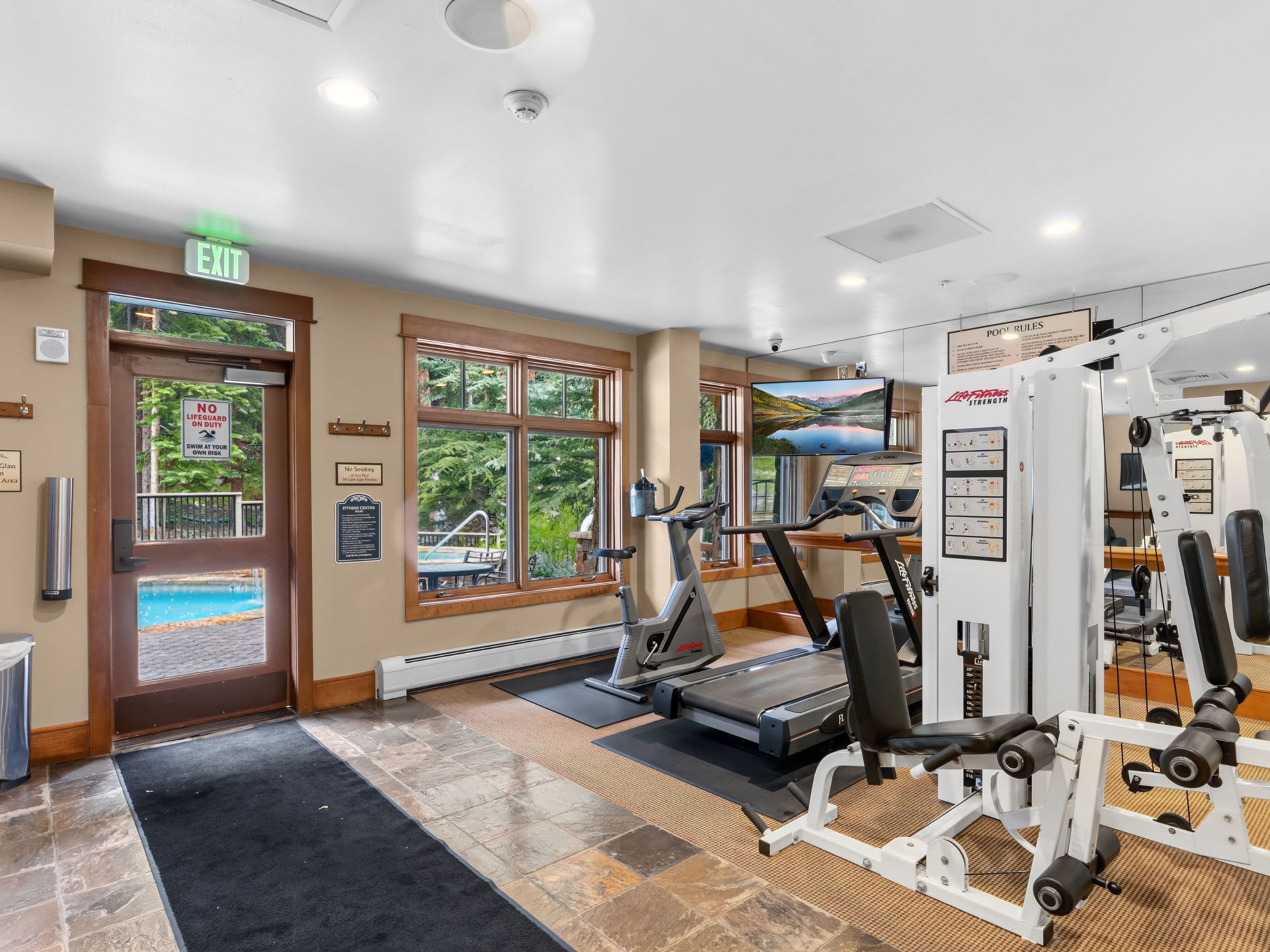 This compact home gym features a variety of fitness equipment, including a stationary bike, a treadmill, and a multi-station strength training machine. The space is finished with a combination of tile and carpeted flooring, and a glass door provides a direct view of an outdoor swimming pool. A wall-mounted television and large mirrors enhance the functionality and perceived size of the room.