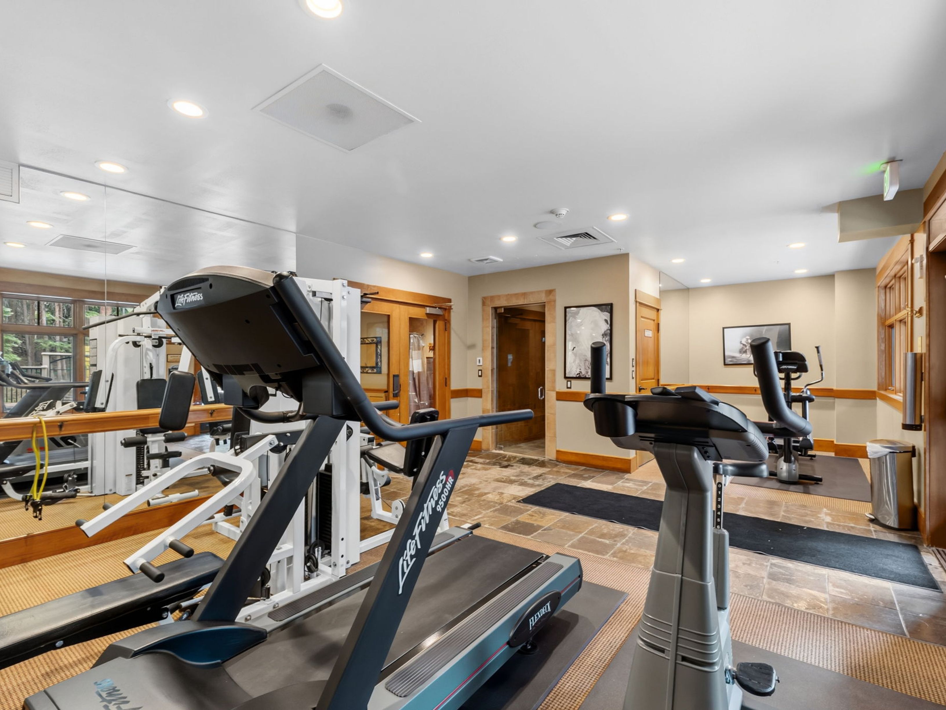 This well-equipped home gym features a Life Fitness treadmill in the foreground and a stationary bike positioned on a rubber mat. The room includes a mirrored wall that enhances the sense of space, along with a multi-station weight machine and tiled flooring. The neutral color palette and recessed lighting create a functional and inviting environment for exercise.