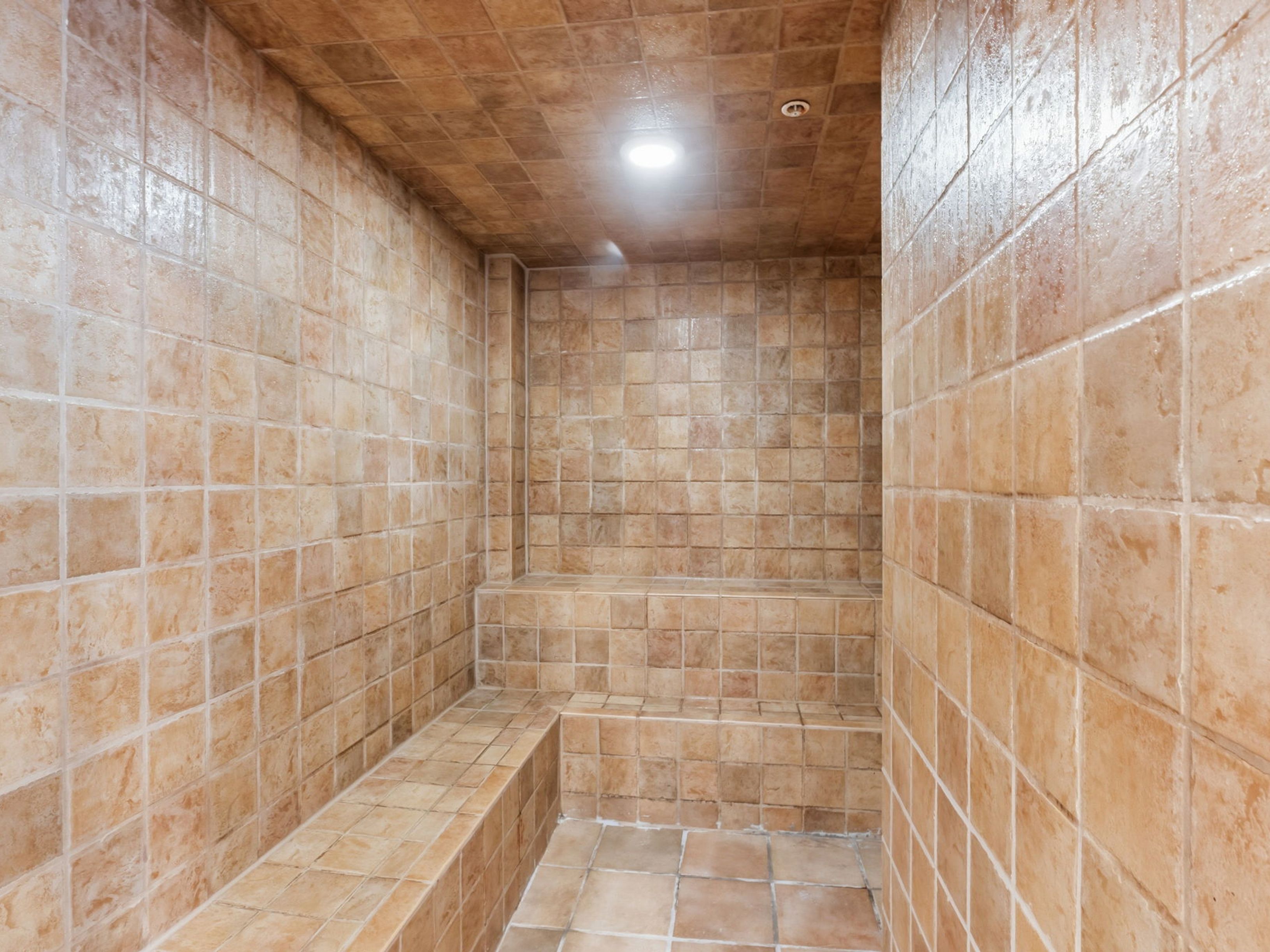 This image showcases a steam room or sauna finished entirely in warm-toned, textured square tiles. The space features built-in tiered bench seating and a recessed ceiling light, creating a compact and enclosed environment. The perspective is centered, highlighting the uniform tile work and the functional layout of the steam room.