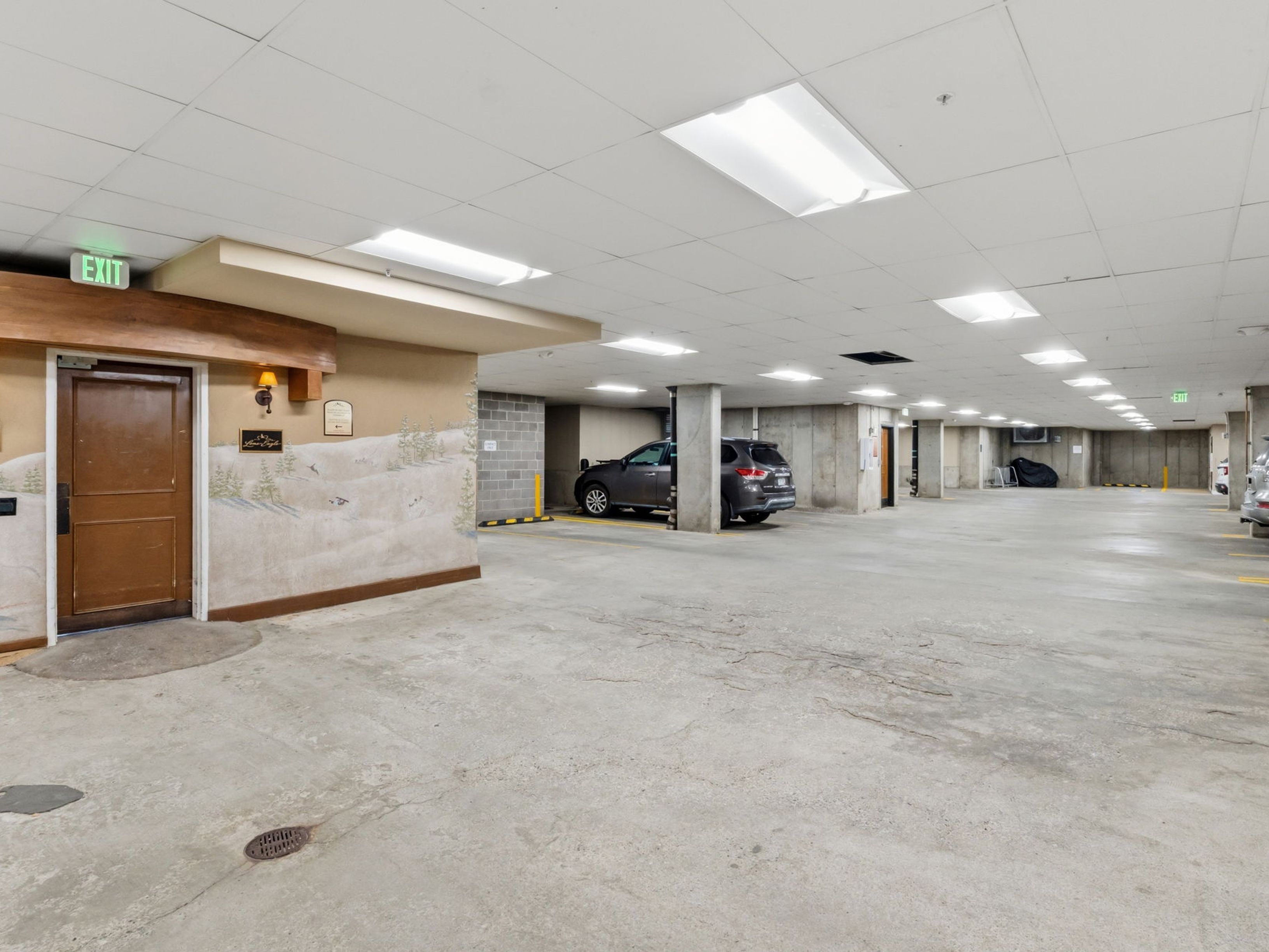This image shows a spacious, well-lit underground parking garage with a concrete floor and exposed structural columns. To the left, a wooden door provides access to the building, accented by a decorative mural of a winter mountain scene on the adjacent wall. The perspective is a wide-angle view looking down the parking lane, highlighting the available parking spaces and the clean, functional layout of the facility.