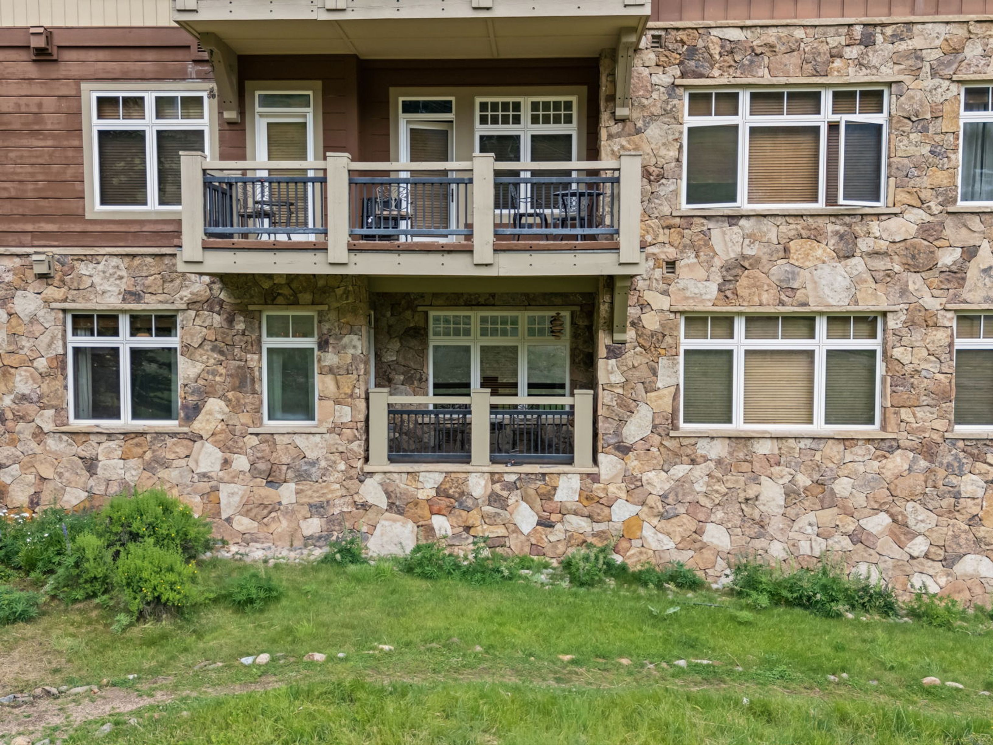 This exterior view captures the rear facade of a multi-story building featuring a rustic aesthetic with stone cladding and dark wood siding. The structure includes a prominent upper-level balcony with a metal railing and a lower-level patio area, both furnished with outdoor seating. Large windows are distributed across the stone wall, and the building is set against a backdrop of green grass and natural landscaping.