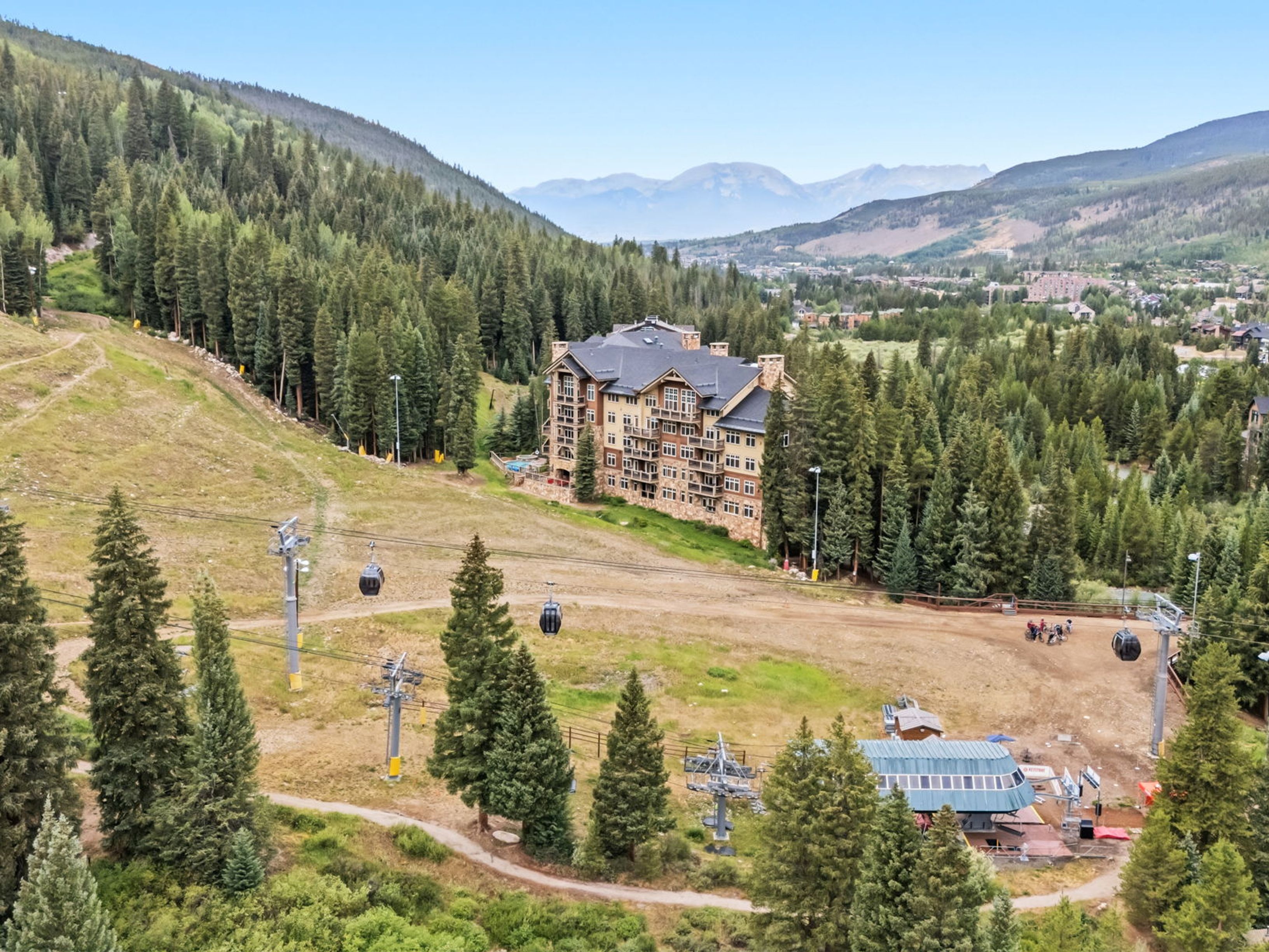 This high-angle aerial view captures a large, multi-story mountain lodge nestled within a dense evergreen forest. The building features a classic alpine architectural style with stone and wood siding, multiple balconies, and a dark pitched roof. In the foreground, a gondola lift system traverses the landscape, highlighting the property's prime location near ski slopes and mountain trails.