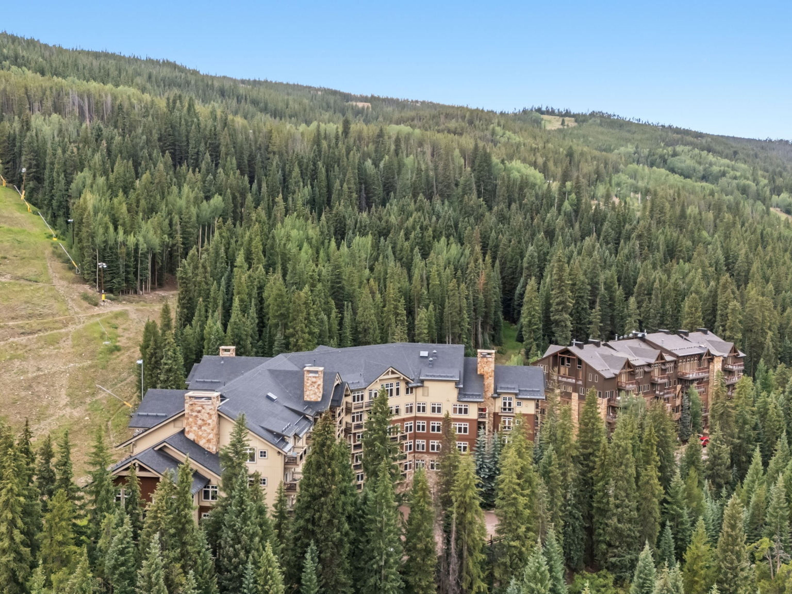 This aerial perspective captures a large, multi-story mountain lodge nestled within a dense forest of evergreen trees. The architecture features a rustic aesthetic with stone-clad chimney breasts, dark shingled roofs, and warm-toned siding that blends into the natural landscape. A ski slope runs along the left side of the property, highlighting its prime location for winter sports enthusiasts.