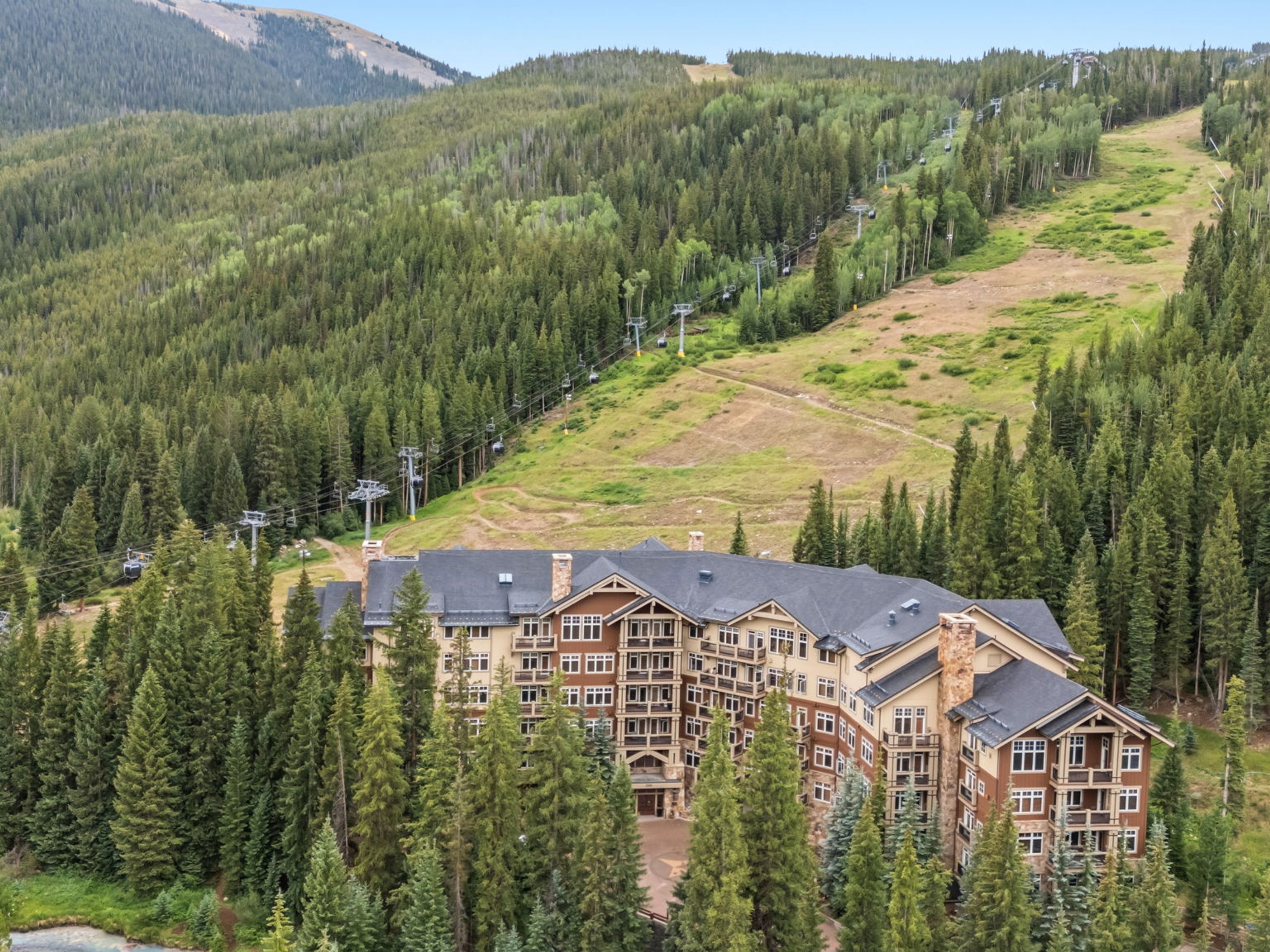 This aerial perspective captures a large, multi-story mountain lodge nestled within a dense evergreen forest at the base of a ski slope. The building features a classic alpine architectural style with a complex roofline, stone accents, and numerous balconies overlooking the surrounding landscape. A ski lift system is visible ascending the mountain behind the property, emphasizing its prime location for winter sports enthusiasts.