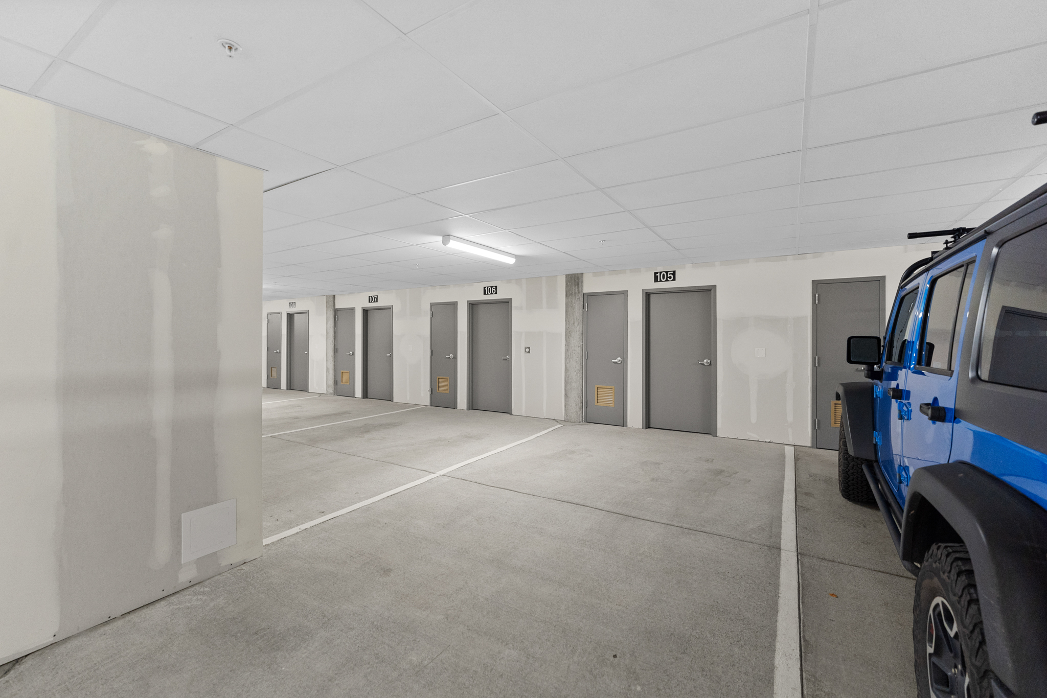 This image shows a clean, modern indoor parking garage featuring a series of numbered storage or utility doors along a white wall. A bright blue Jeep is parked on the right side, adding a pop of color to the neutral concrete and drywall aesthetic. The perspective is from the entrance of the parking bay, looking down the row of doors under bright, overhead fluorescent lighting.