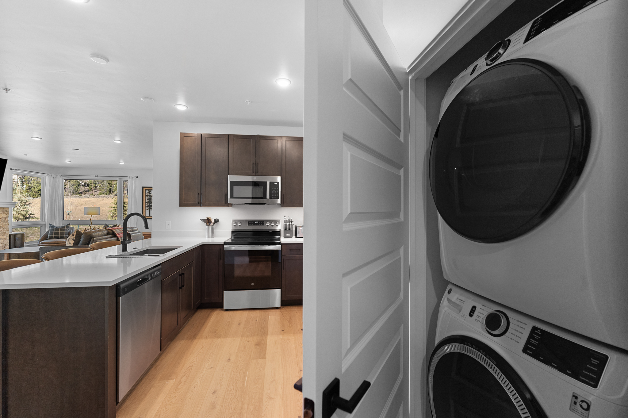 This image captures a view from a laundry closet looking out into a modern, open-concept kitchen. The kitchen features dark wood cabinetry, stainless steel appliances, and a sleek white quartz island countertop, creating a sophisticated and functional space. The perspective highlights the seamless transition between the utility area and the main living space, which includes a glimpse of a bright, window-filled living room in the background.