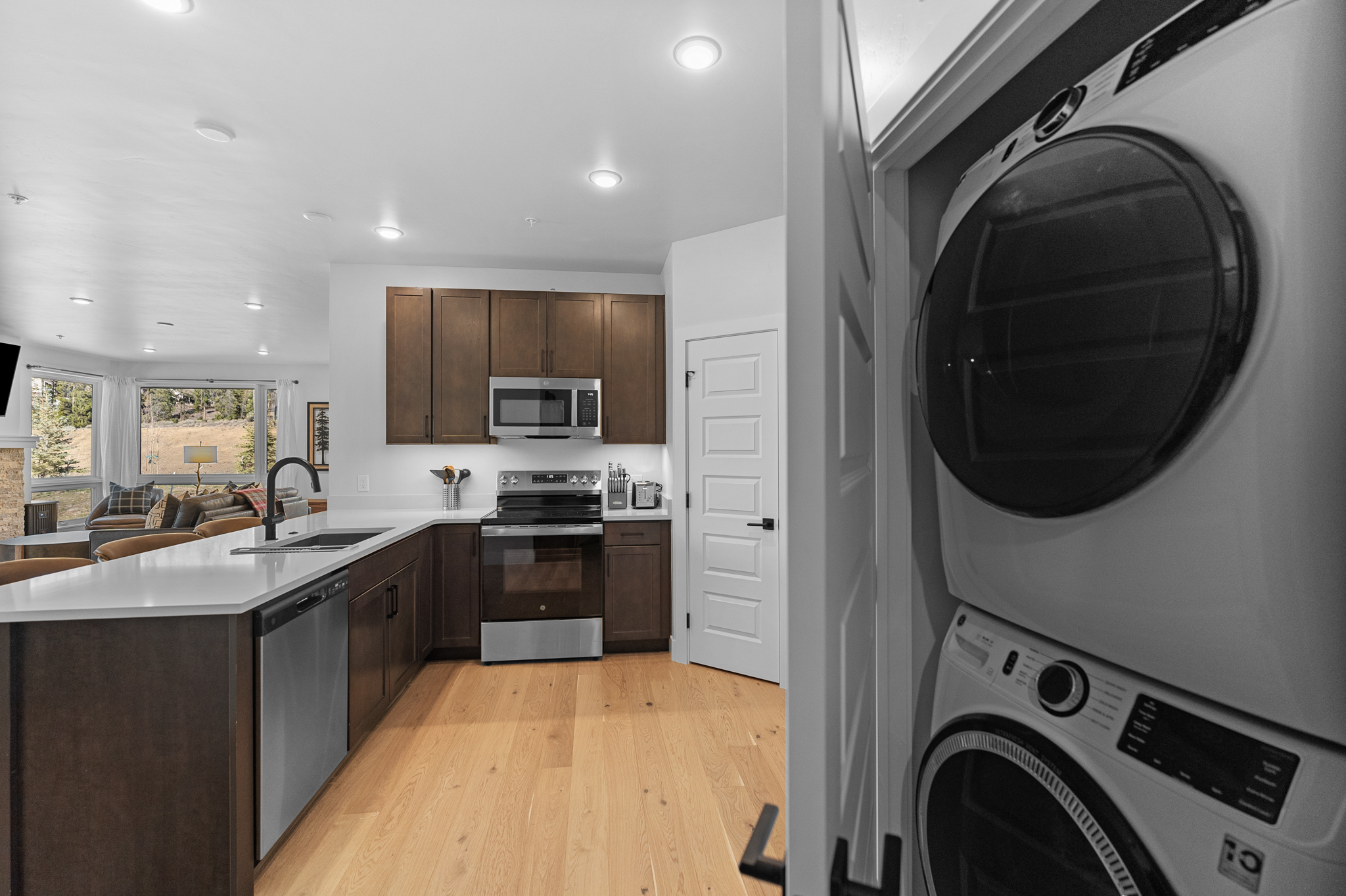 This modern kitchen features dark wood cabinetry, stainless steel appliances, and a sleek white quartz countertop island that flows into an open-concept living area. The perspective is captured from the doorway, highlighting a stacked washer and dryer unit in the foreground, which transitions into the warm, light-wood floored kitchen space. Large windows in the background provide a bright, scenic view, creating an inviting and functional atmosphere.