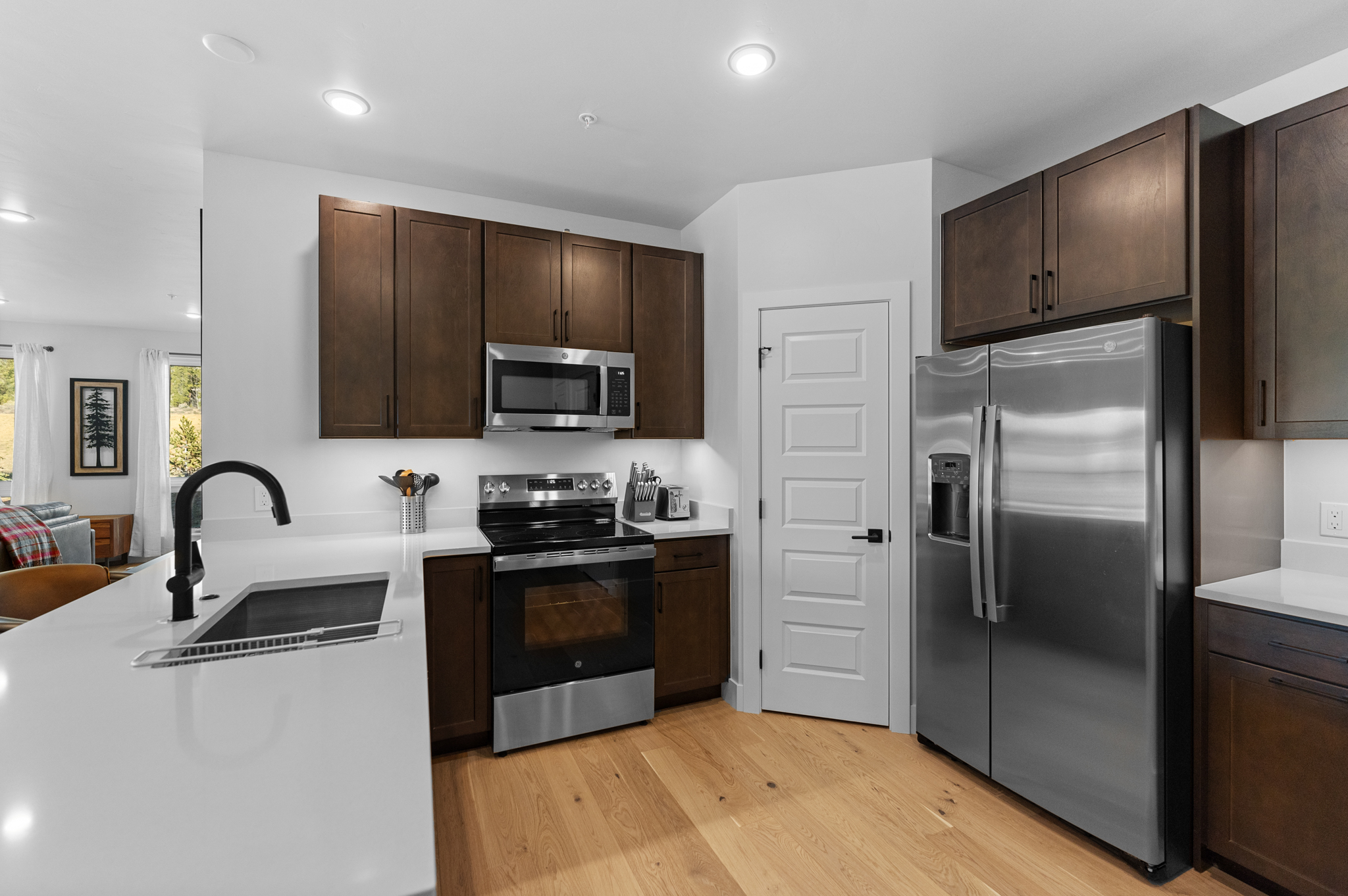 This modern kitchen features rich, dark wood cabinetry contrasted by sleek white quartz countertops and stainless steel appliances. The space includes a functional island with a black faucet, a built-in microwave, and a full-sized refrigerator, all set against light hardwood flooring. The clean lines and neutral color palette create an inviting and contemporary atmosphere perfect for culinary activities.