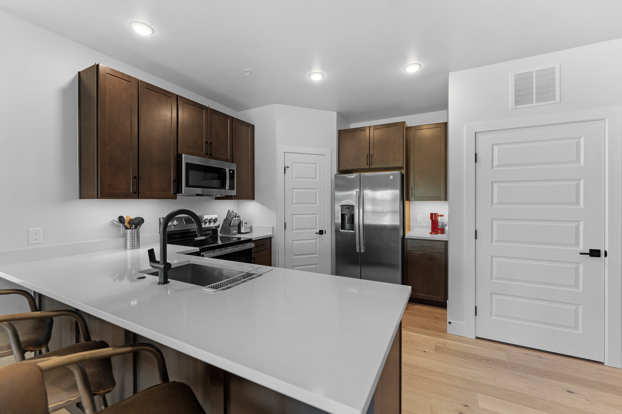 This modern kitchen features dark wood cabinetry contrasted by sleek white quartz countertops and a large island with bar seating. The space is equipped with stainless steel appliances, including a built-in microwave and a side-by-side refrigerator, all illuminated by recessed ceiling lighting. The open-concept design flows seamlessly into the surrounding area, creating a clean, functional, and inviting atmosphere.