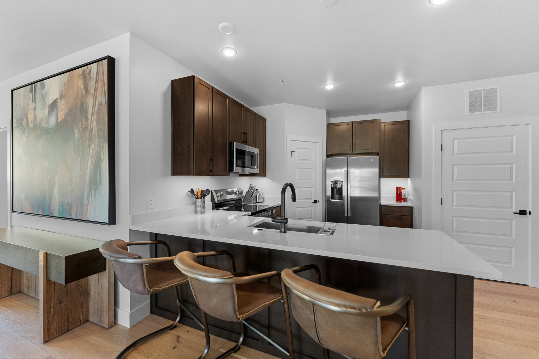 This modern kitchen features dark wood cabinetry contrasted by sleek white quartz countertops and a breakfast bar with three leather-upholstered stools. The space is equipped with stainless steel appliances, including a built-in microwave and a refrigerator, and is illuminated by recessed lighting. A large abstract painting on the adjacent wall adds a sophisticated touch to the open-concept layout.