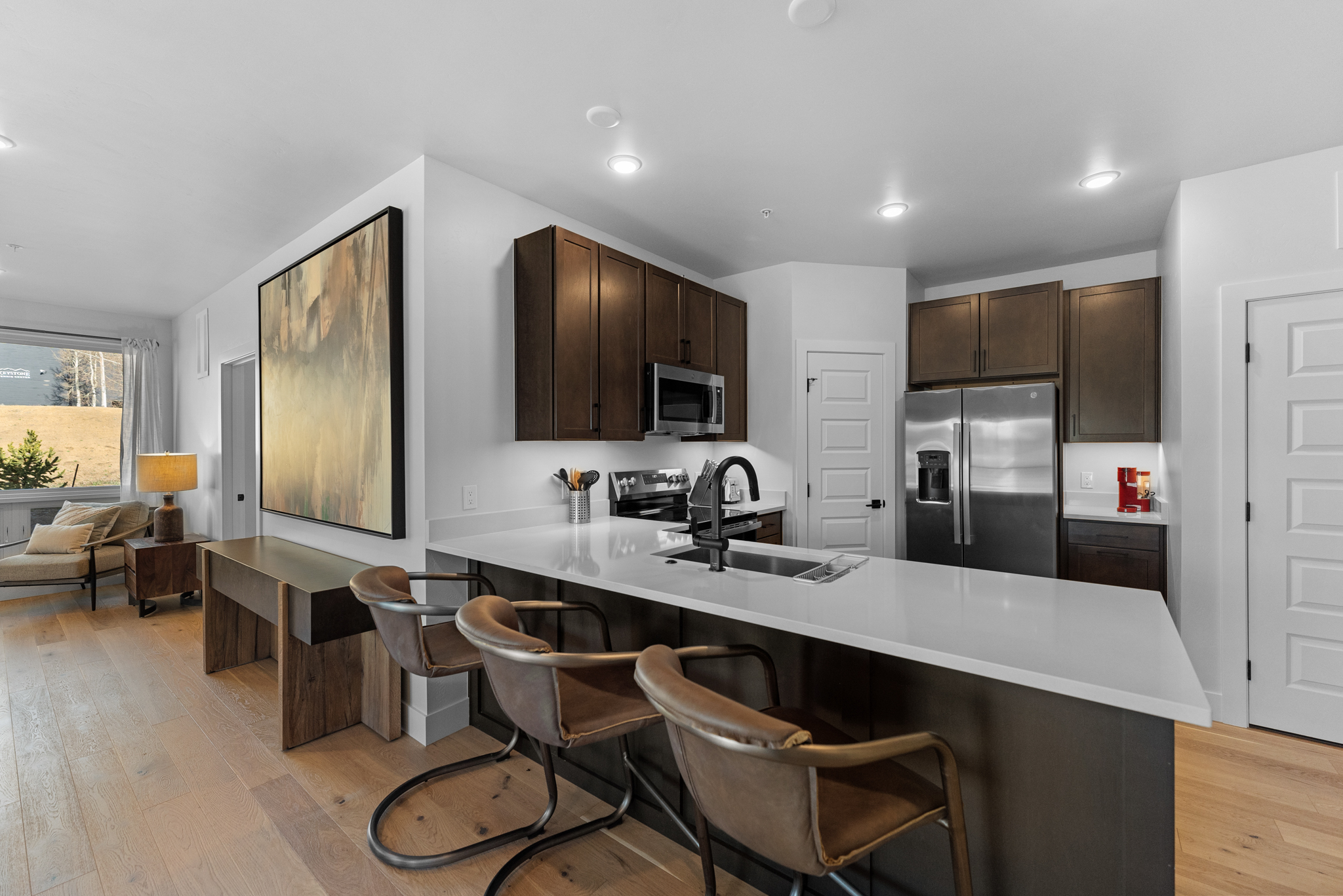 This modern kitchen features dark wood cabinetry, sleek white quartz countertops, and a functional island with three leather-upholstered bar stools. The open-concept design seamlessly transitions into a cozy living area, highlighted by a large abstract painting and light hardwood flooring throughout. The space is well-lit with recessed lighting and offers a clean, sophisticated aesthetic perfect for contemporary living.