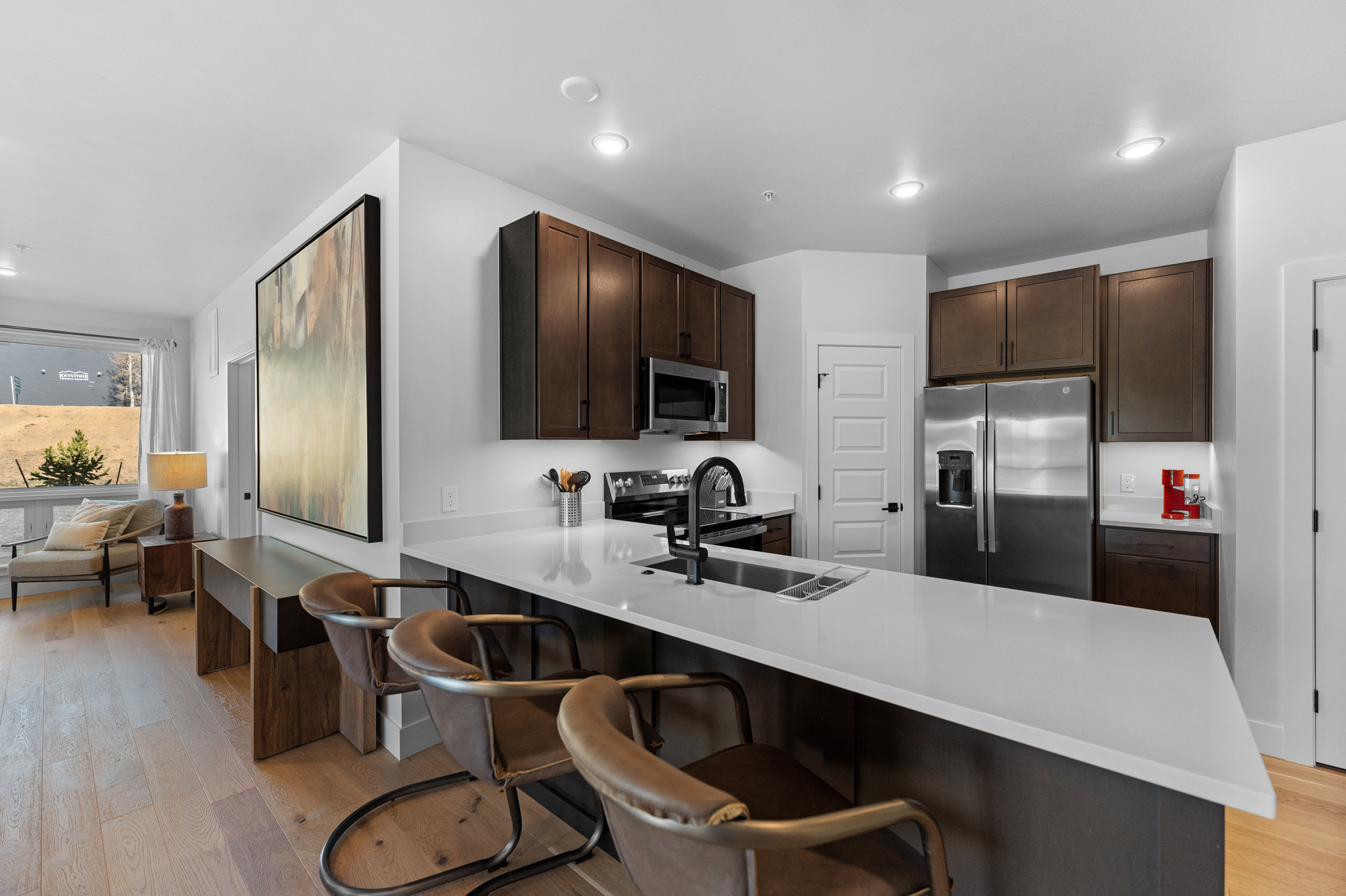 This modern, open-concept kitchen features dark wood cabinetry, sleek white quartz countertops, and high-end stainless steel appliances. A spacious island with three leather-upholstered bar stools serves as a central gathering point, while the adjacent living area is visible through a wide opening. The space is bright and inviting, characterized by clean lines, contemporary finishes, and a seamless transition between the culinary and relaxation zones.