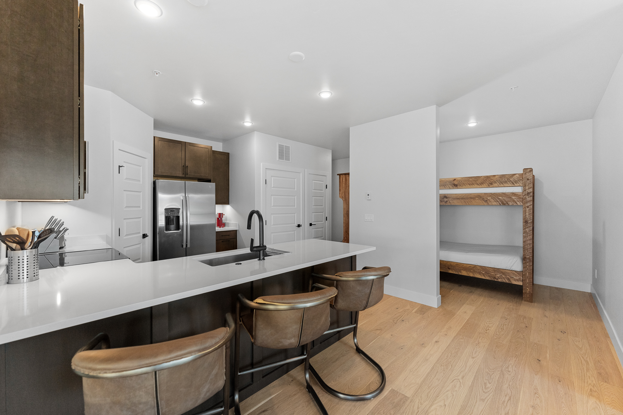 This modern, open-concept kitchen features dark cabinetry contrasted by crisp white countertops and light hardwood flooring. A central island with bar seating serves as a functional focal point, while a rustic wooden bunk bed is tucked into an adjacent alcove, maximizing space efficiency. The clean lines and neutral color palette create a contemporary and inviting atmosphere suitable for a vacation rental or compact living space.