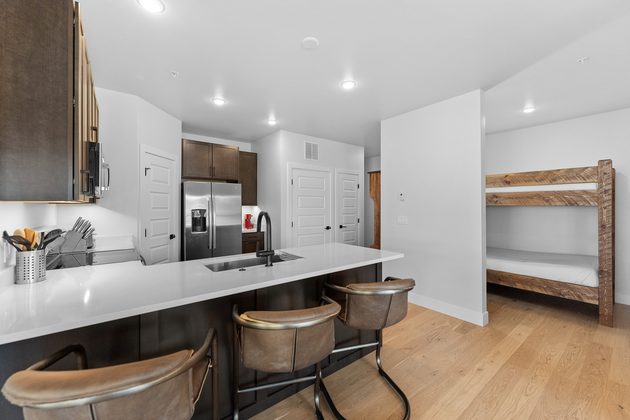 This modern, open-concept kitchen features dark wood cabinetry, sleek white quartz countertops, and a central island with leather-upholstered bar stools. The space seamlessly integrates a functional cooking area with a cozy, built-in wooden bunk bed nook, highlighting a clever use of space. The light hardwood flooring and recessed lighting create a bright, inviting atmosphere perfect for contemporary living.