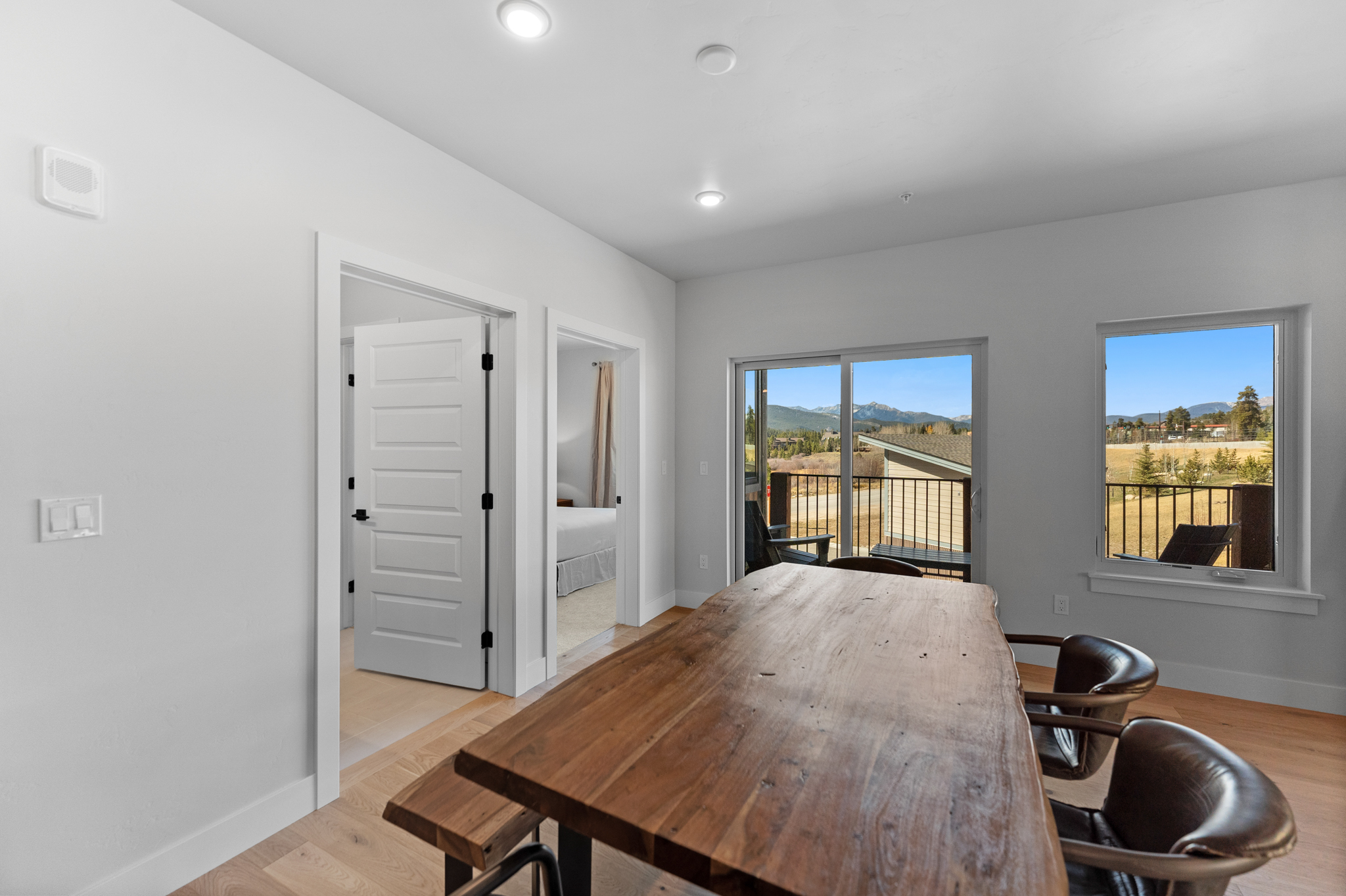 This bright and airy dining room features a rustic, live-edge wooden table paired with leather chairs, creating a warm and inviting atmosphere. The space is highlighted by light hardwood flooring, clean white walls, and a sliding glass door that provides a scenic view of the surrounding mountains. The perspective captures the transition from the indoor dining area to the outdoor balcony, emphasizing the home's connection to the natural landscape.