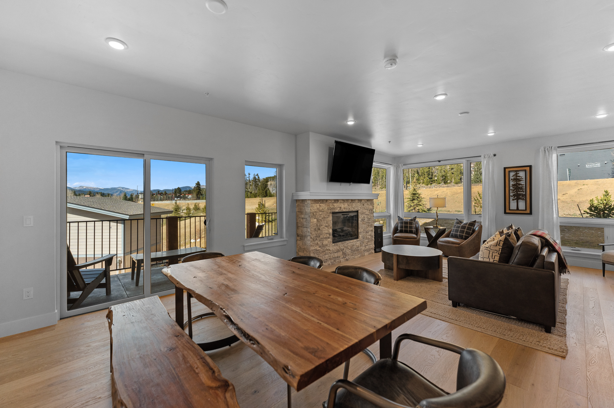 This open-concept living and dining area features a rustic, natural-edge wood dining table and a cozy living space anchored by a stone fireplace with a mounted television. Large sliding glass doors and expansive windows flood the room with natural light, showcasing scenic mountain views and a private balcony. The interior design blends modern comfort with mountain-lodge aesthetics, utilizing warm wood tones, leather furniture, and neutral textures to create an inviting atmosphere.