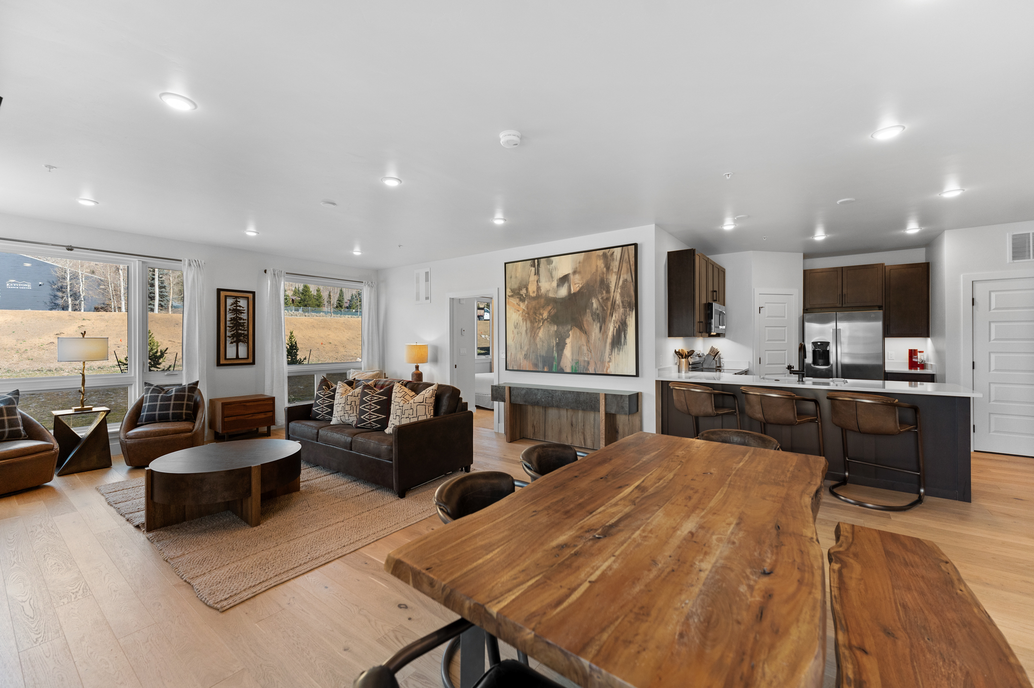 This open-concept living area features a warm, rustic-modern aesthetic with light hardwood flooring and a large, live-edge wooden dining table in the foreground. The space flows seamlessly into a cozy living room with a dark leather sofa, a unique oval coffee table, and large windows that provide ample natural light and scenic views. A sophisticated kitchen with a breakfast bar and dark cabinetry completes the space, making it ideal for both relaxation and entertaining.