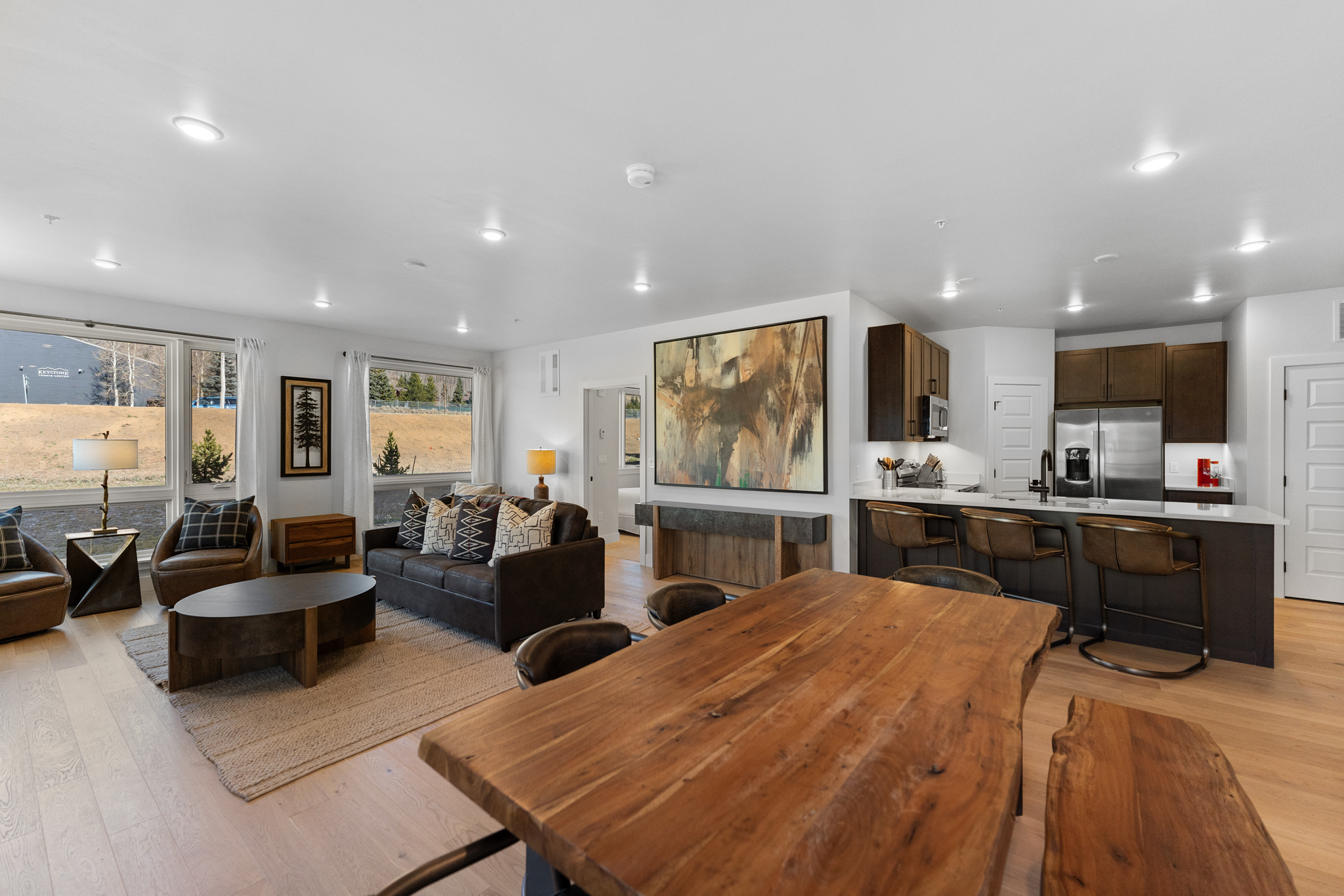 This open-concept living space features a warm, rustic-modern aesthetic with light hardwood floors and a large, live-edge wooden dining table in the foreground. The living area is anchored by a dark leather sofa, a matching armchair, and a unique oval coffee table, all set against a backdrop of large windows that provide natural light and scenic views. A prominent abstract painting hangs above a console table, bridging the transition between the cozy seating area and the adjacent kitchen, which is equipped with dark cabinetry and bar seating.
