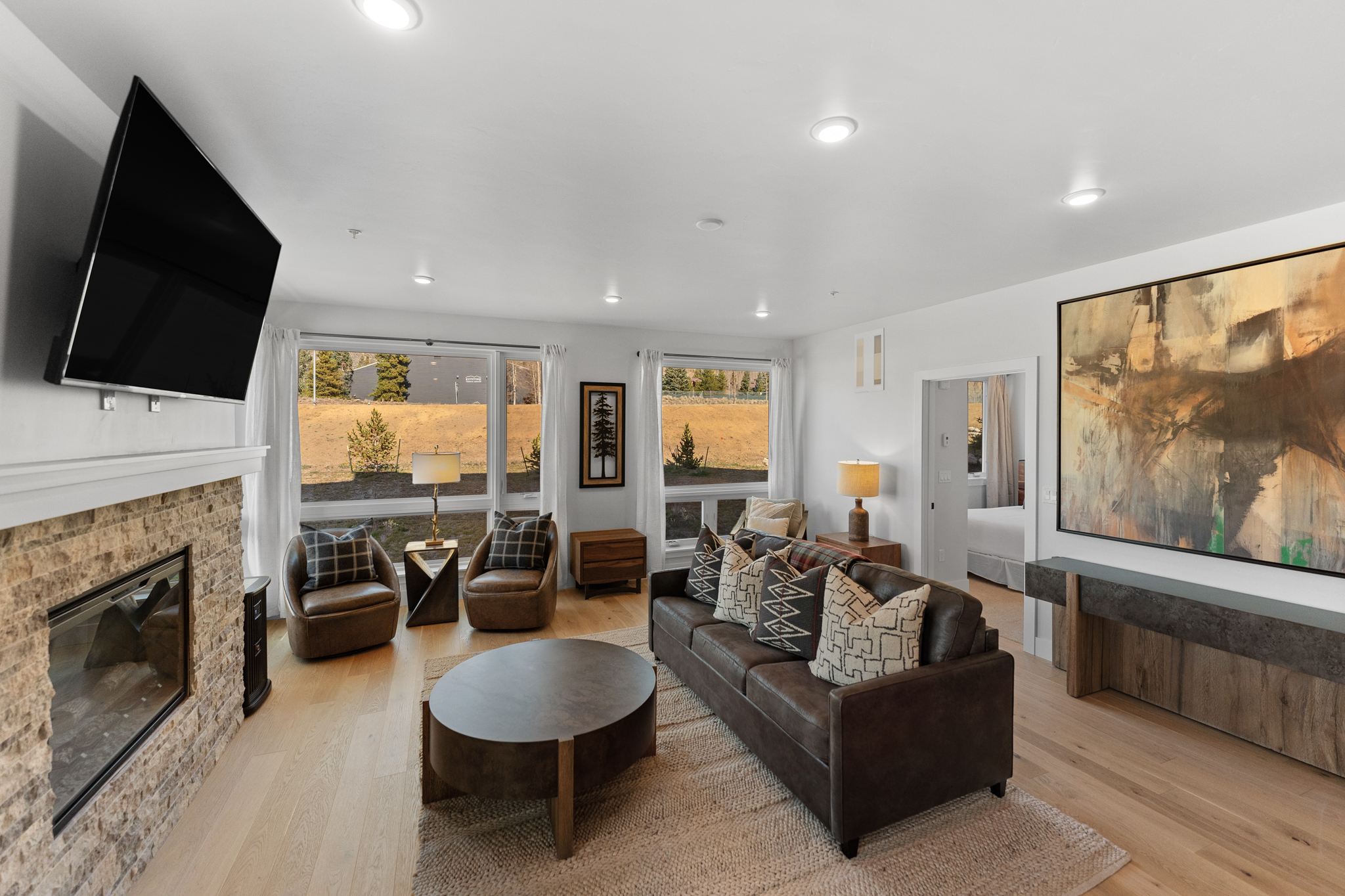 This inviting living room features a warm, rustic-modern aesthetic with a stone-faced fireplace, a dark leather sofa, and matching armchairs arranged on a textured area rug. Large windows provide ample natural light and scenic views of the outdoors, while a prominent abstract painting adds a sophisticated focal point to the space. The open-concept layout flows seamlessly into an adjacent bedroom, creating a comfortable and cohesive living environment.