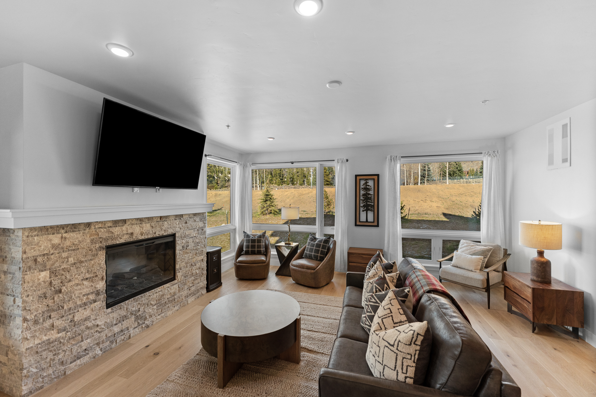 This inviting living room features a prominent stone fireplace with a mounted television, creating a cozy focal point. The space is furnished with a dark leather sofa, patterned throw pillows, and two leather armchairs, all arranged on a textured area rug over light hardwood floors. Large windows provide ample natural light and scenic views of the surrounding landscape, contributing to a warm, rustic-modern aesthetic.