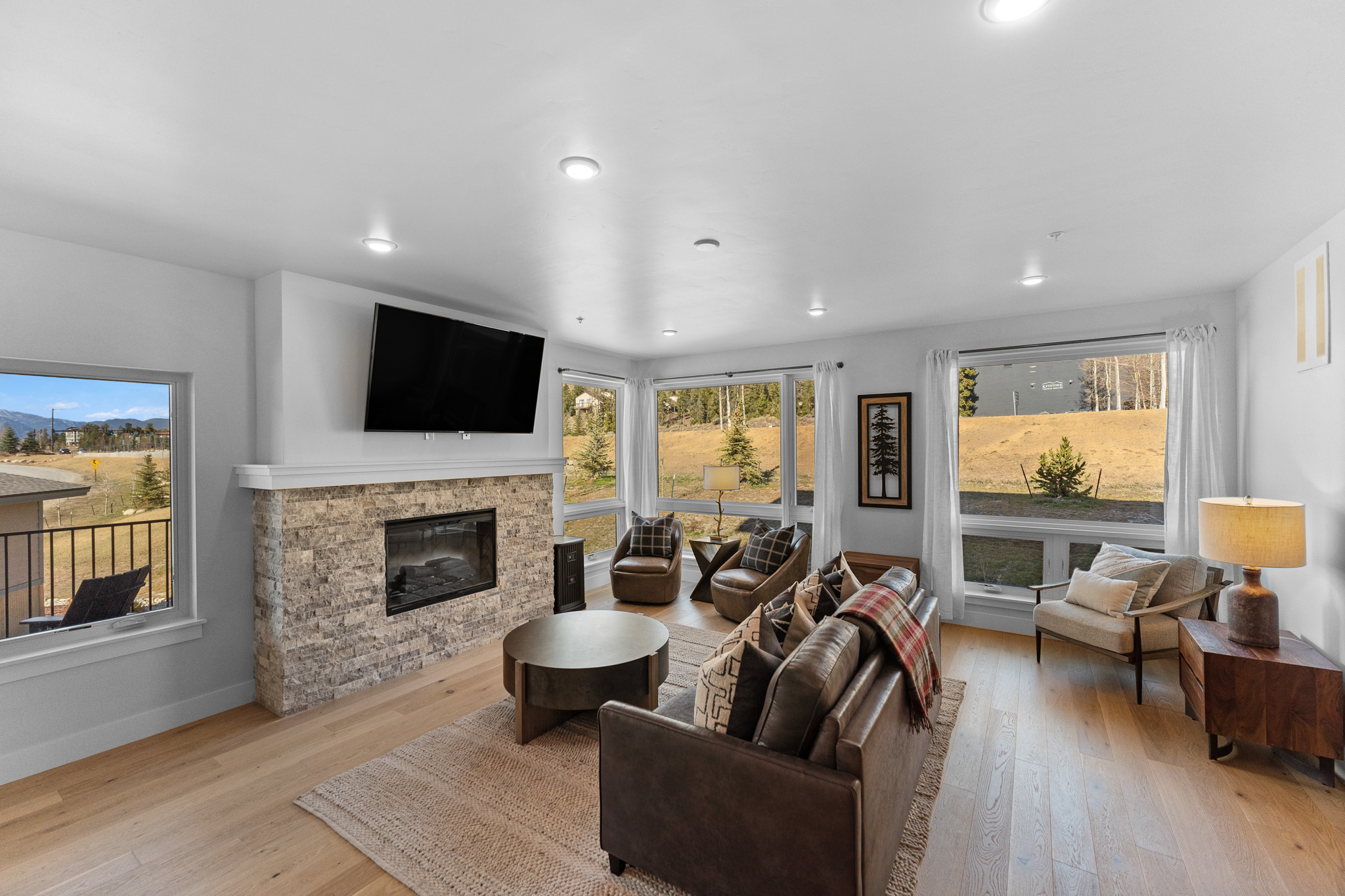 This inviting living room features a warm, rustic-modern aesthetic with a prominent stone-clad fireplace and a mounted television as the focal point. The space is furnished with a comfortable leather sofa, a round coffee table, and several accent chairs, all arranged on a textured area rug over light hardwood floors. Large windows flood the room with natural light and offer expansive views of the surrounding landscape, creating a bright and airy atmosphere.