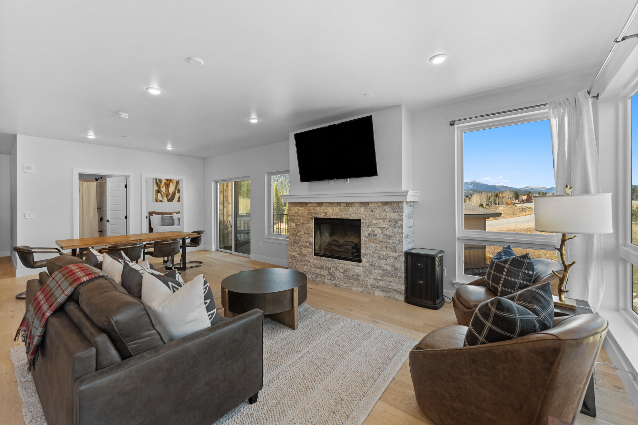 This spacious, open-concept living room features a cozy stone fireplace as its focal point, topped with a large mounted television. The room is furnished with a comfortable leather sofa and matching armchairs, all arranged on a textured area rug, while large windows provide abundant natural light and scenic mountain views. The neutral color palette and warm wood flooring create an inviting, modern mountain-lodge aesthetic.