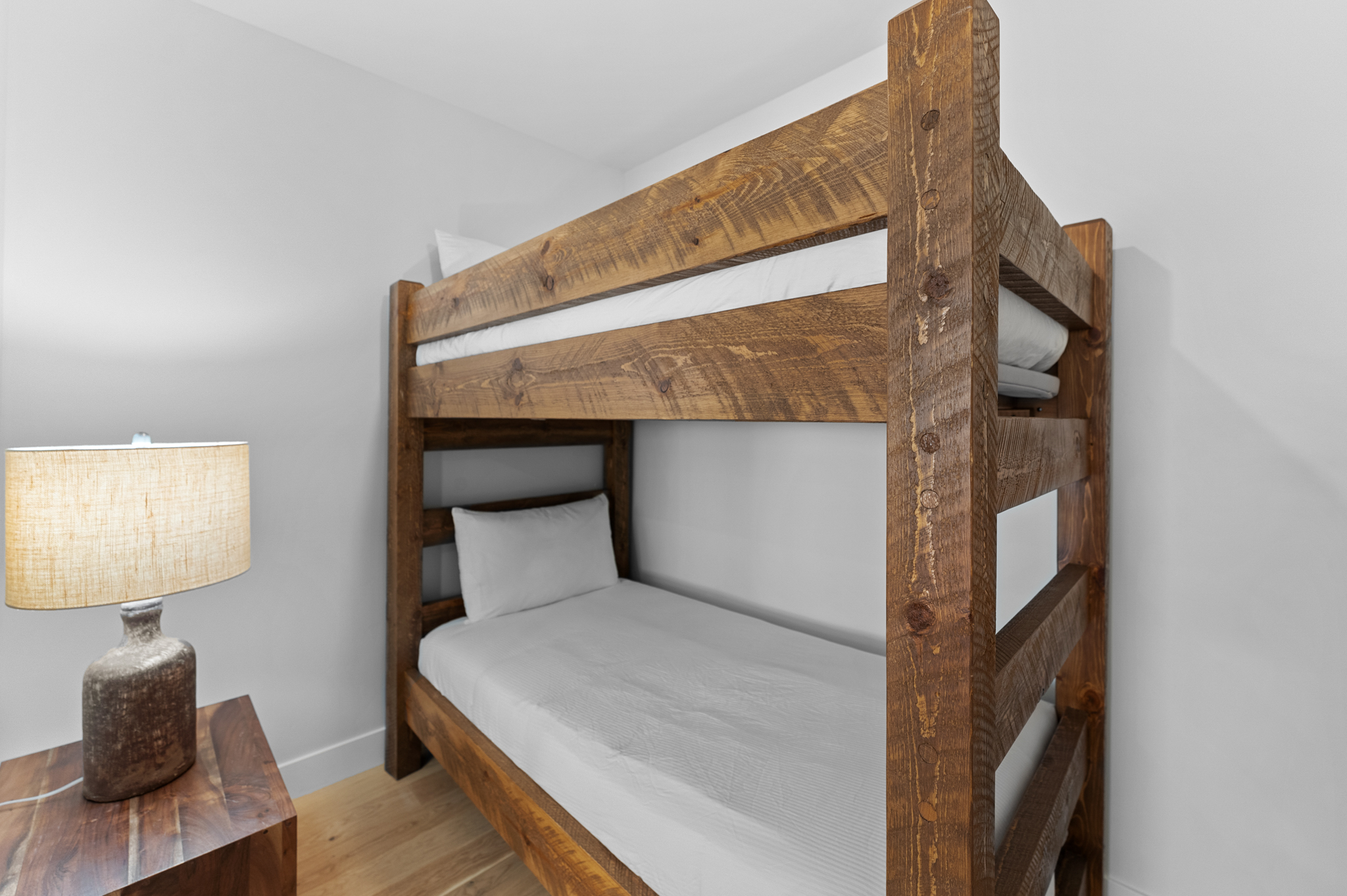 This cozy bedroom features a rustic, heavy-timber bunk bed set against a neutral white wall, creating a warm and inviting atmosphere. To the left, a wooden nightstand holds a textured, earth-toned lamp that provides soft, ambient lighting. The room is finished with light-colored hardwood flooring, emphasizing a clean and modern aesthetic suitable for a guest room or children's space.