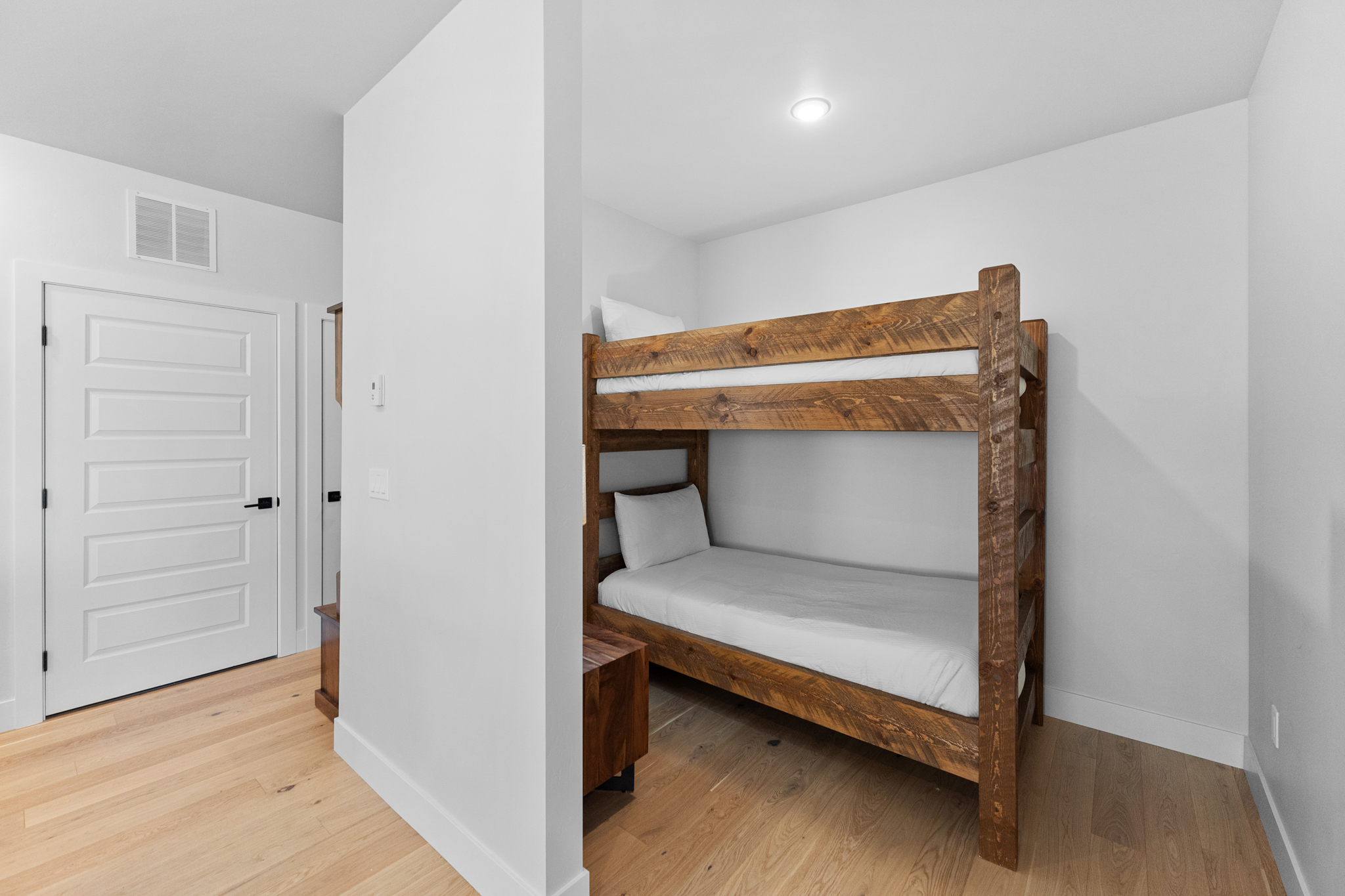 This cozy bedroom alcove features a rustic, dark-stained wooden bunk bed set against a clean, white-walled backdrop. The space is accented by light-toned hardwood flooring and a minimalist aesthetic, creating a warm and inviting atmosphere. The perspective captures the bunk bed tucked into a nook, adjacent to a white interior door, highlighting an efficient use of space.