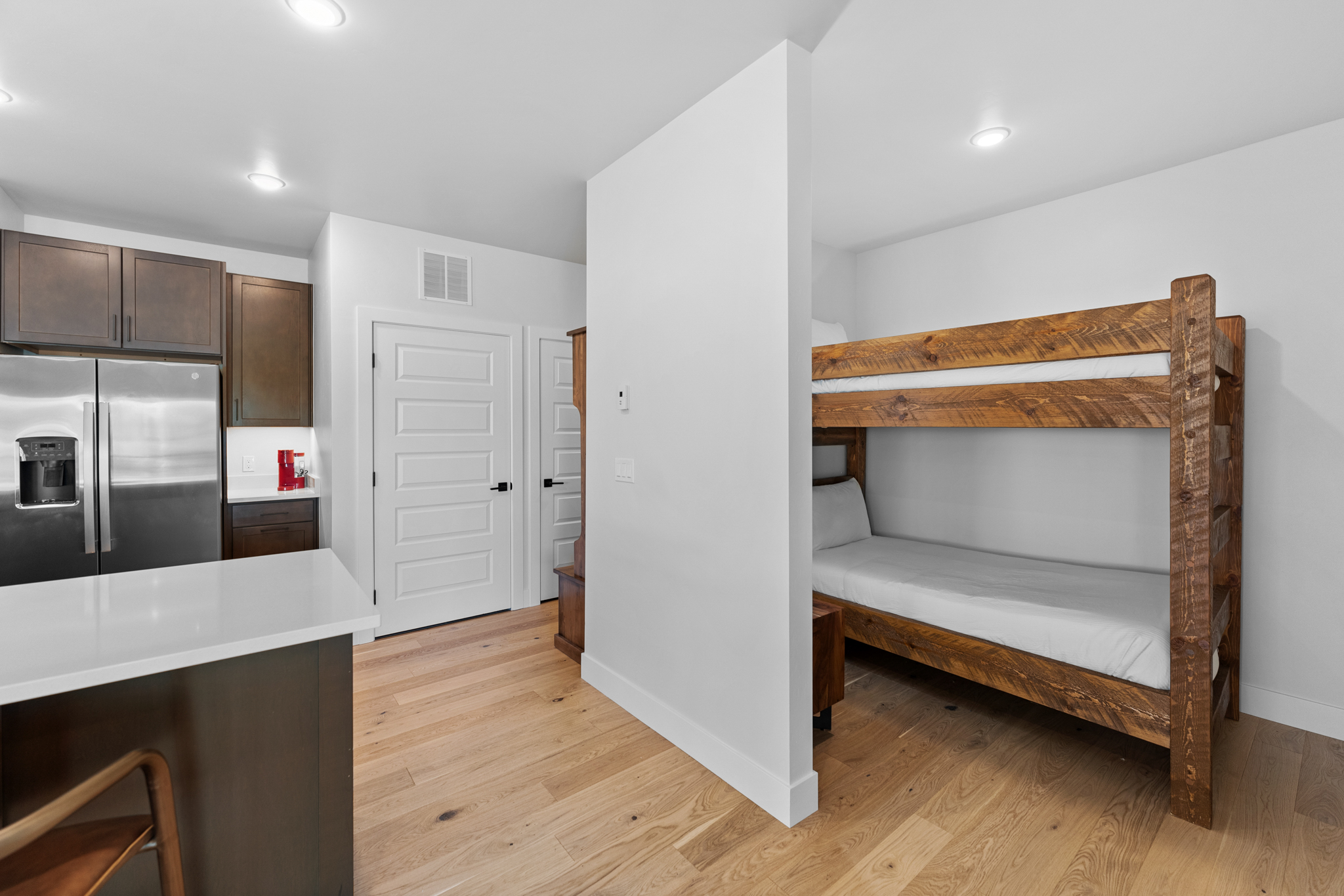 This interior shot captures a compact, modern living space featuring a rustic wooden bunk bed tucked behind a partial partition wall. To the left, a sleek kitchen area with stainless steel appliances and dark cabinetry is visible, creating a functional open-concept layout. The light wood flooring and neutral color palette give the room a clean, contemporary, and efficient feel, ideal for a vacation rental or guest suite.