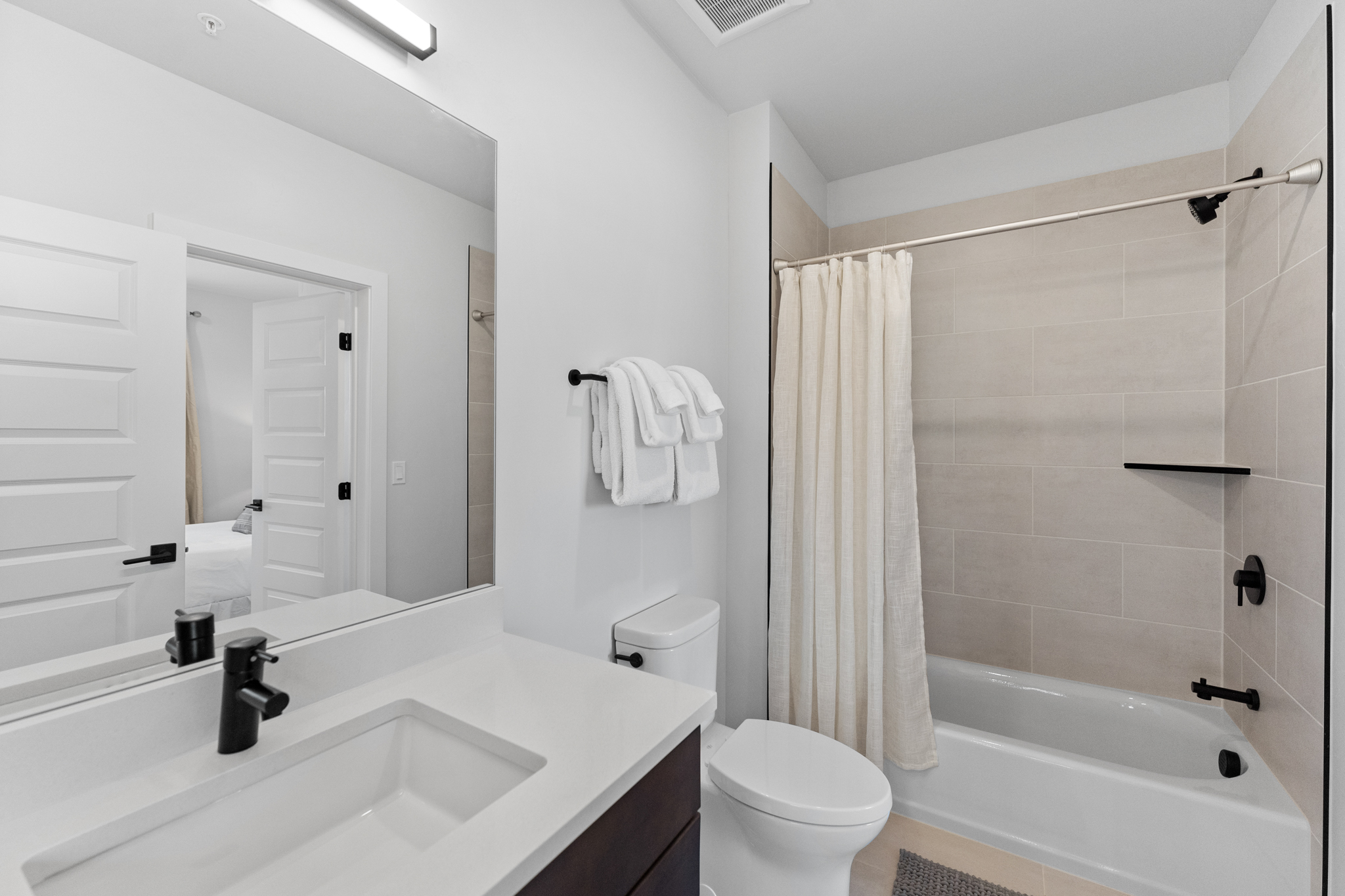 This modern, clean-lined bathroom features a sleek vanity with a white countertop, a large wall-mounted mirror, and a shower-tub combination with neutral-toned tile work. The space is accented with contemporary matte black fixtures, including the faucet, shower head, and towel hardware, creating a sophisticated and cohesive aesthetic. The bright, neutral color palette and efficient layout make this a functional and stylish space.