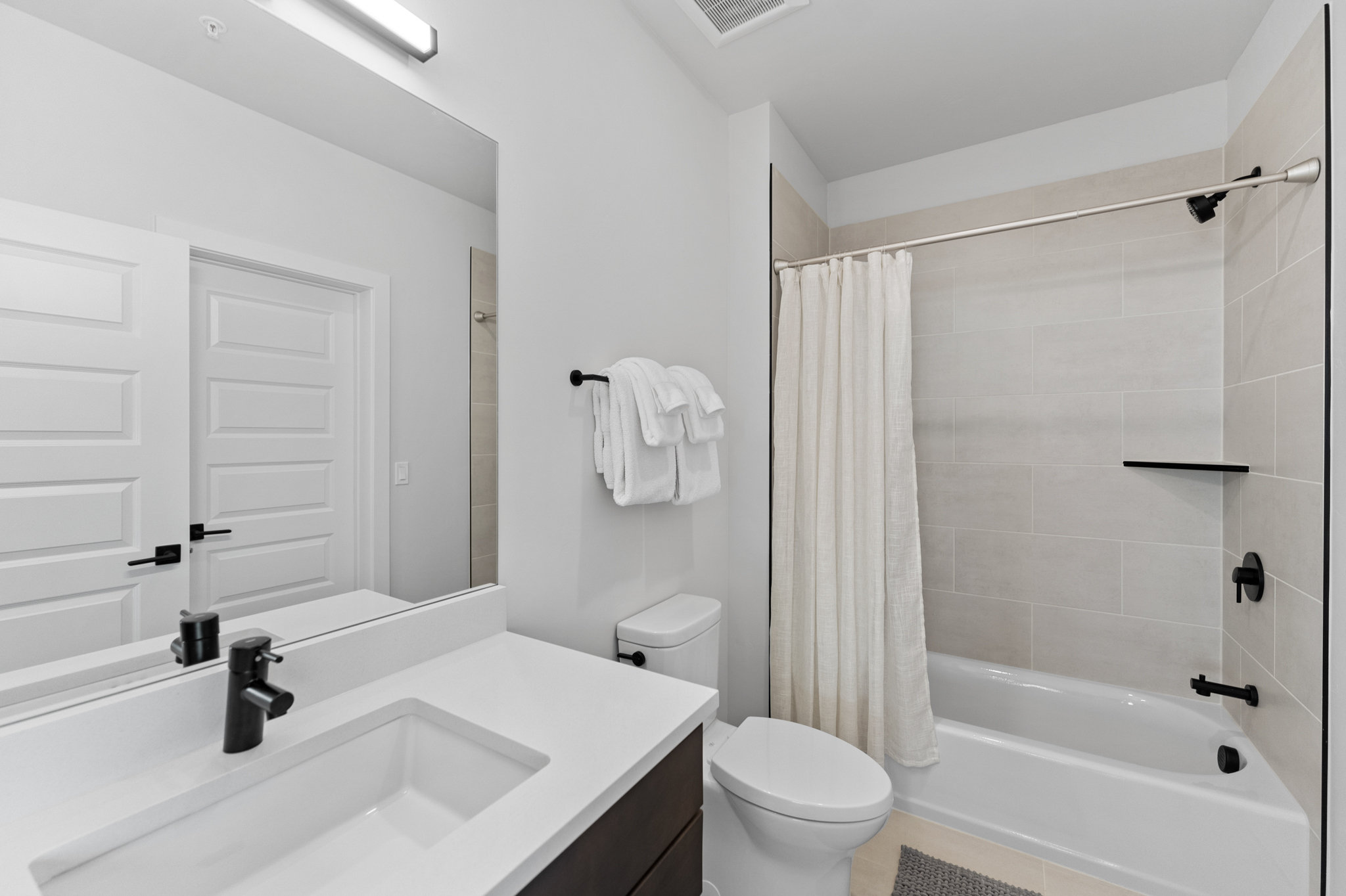 This modern, clean-lined bathroom features a sleek vanity with a white countertop, a black faucet, and a large mirror that enhances the sense of space. The shower-tub combination is finished with light-toned wall tiles, a black shower head, and a neutral shower curtain, creating a bright and functional aesthetic. The overall design is contemporary and minimalist, making it an appealing space for guests.