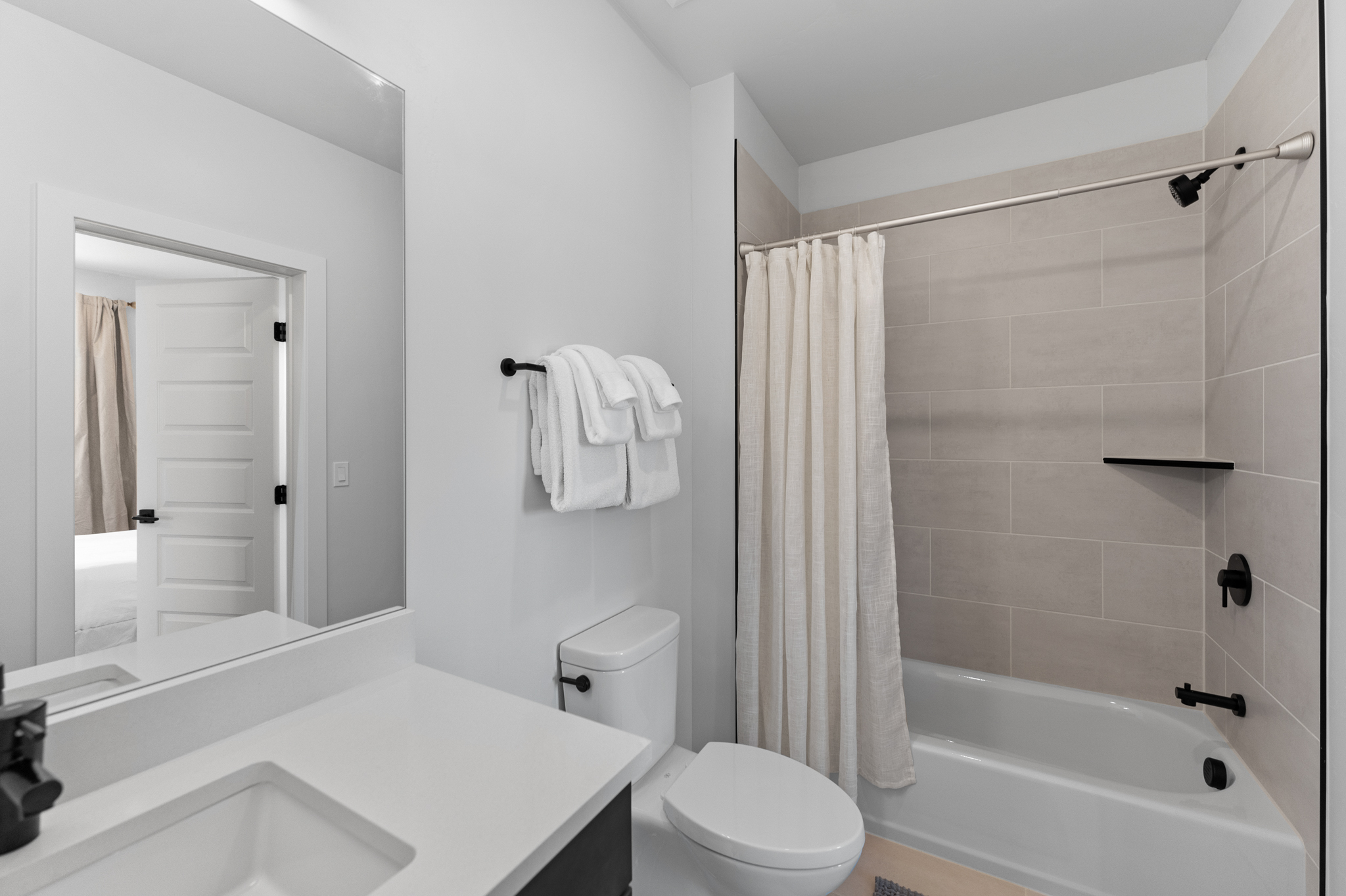 This clean and modern bathroom features a neutral color palette with light gray wall tiles in the shower area and a white vanity. The space includes a shower-tub combination with matte black fixtures, a wall-mounted towel rack, and a large mirror reflecting the adjacent bedroom doorway. The overall aesthetic is minimalist and functional, providing a bright and well-maintained appearance.