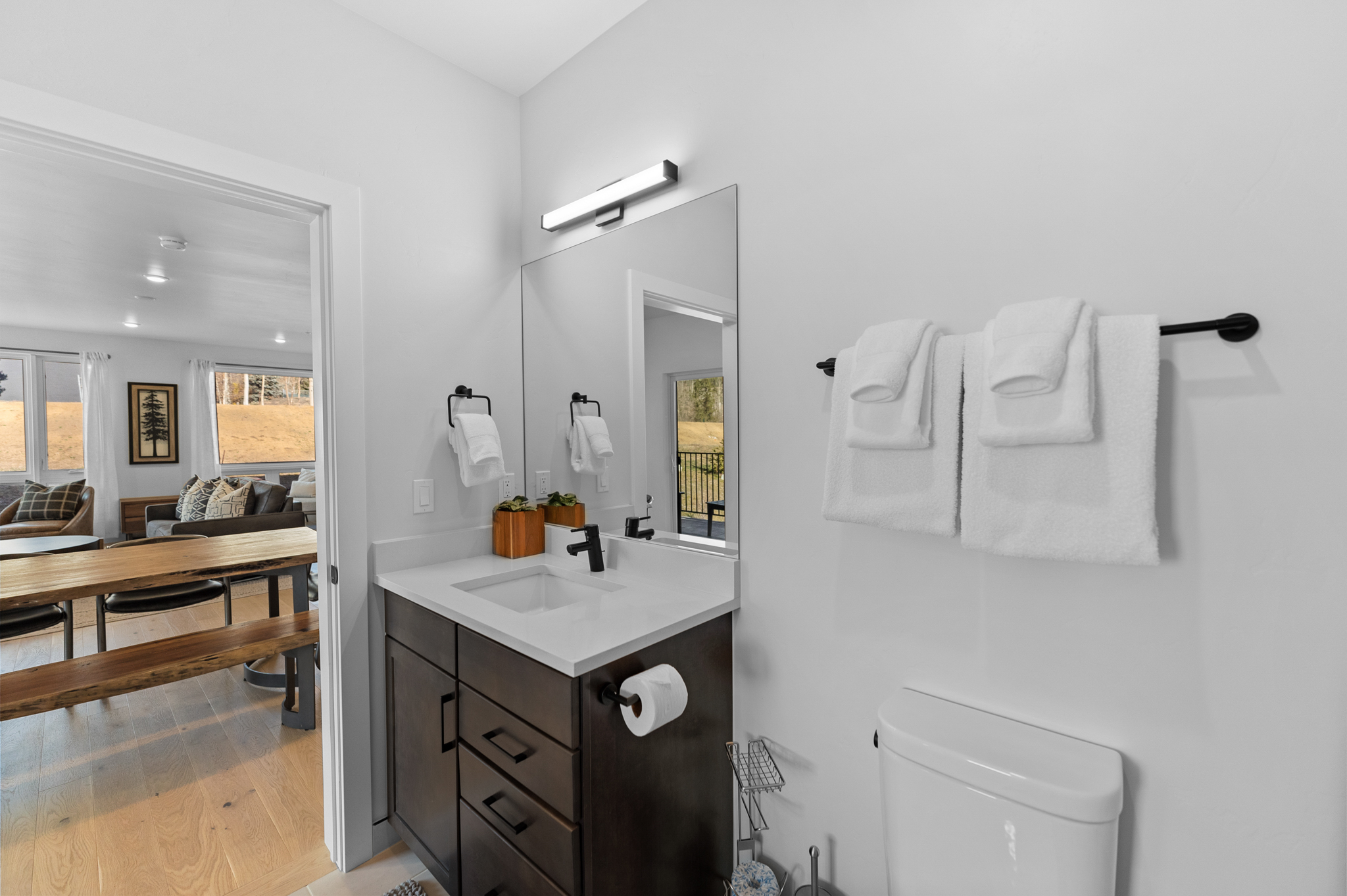 This modern bathroom features a dark wood vanity with a white quartz countertop, a sleek black faucet, and a large rectangular mirror. The space is bright and clean, with white towels hanging on a black rack and a view through the doorway into an adjacent dining area. The minimalist design and neutral color palette create a polished and contemporary aesthetic.
