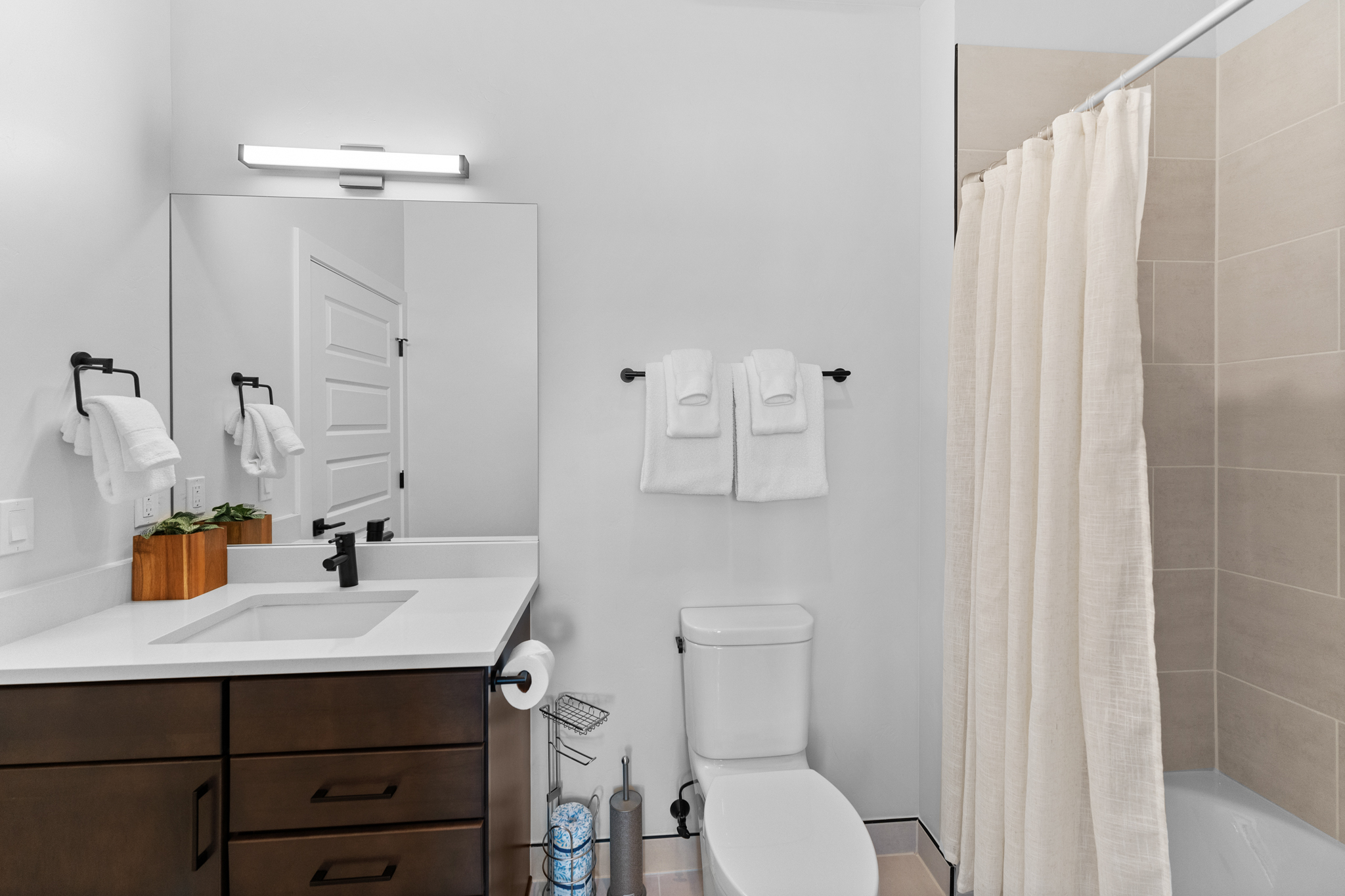 This clean and modern bathroom features a dark wood vanity with a white quartz countertop and a sleek black faucet. The space includes a wall-mounted mirror, a minimalist light fixture, and a shower-tub combination with neutral-toned tiling and a light-colored curtain. The overall aesthetic is contemporary and functional, making it an appealing space for guests.