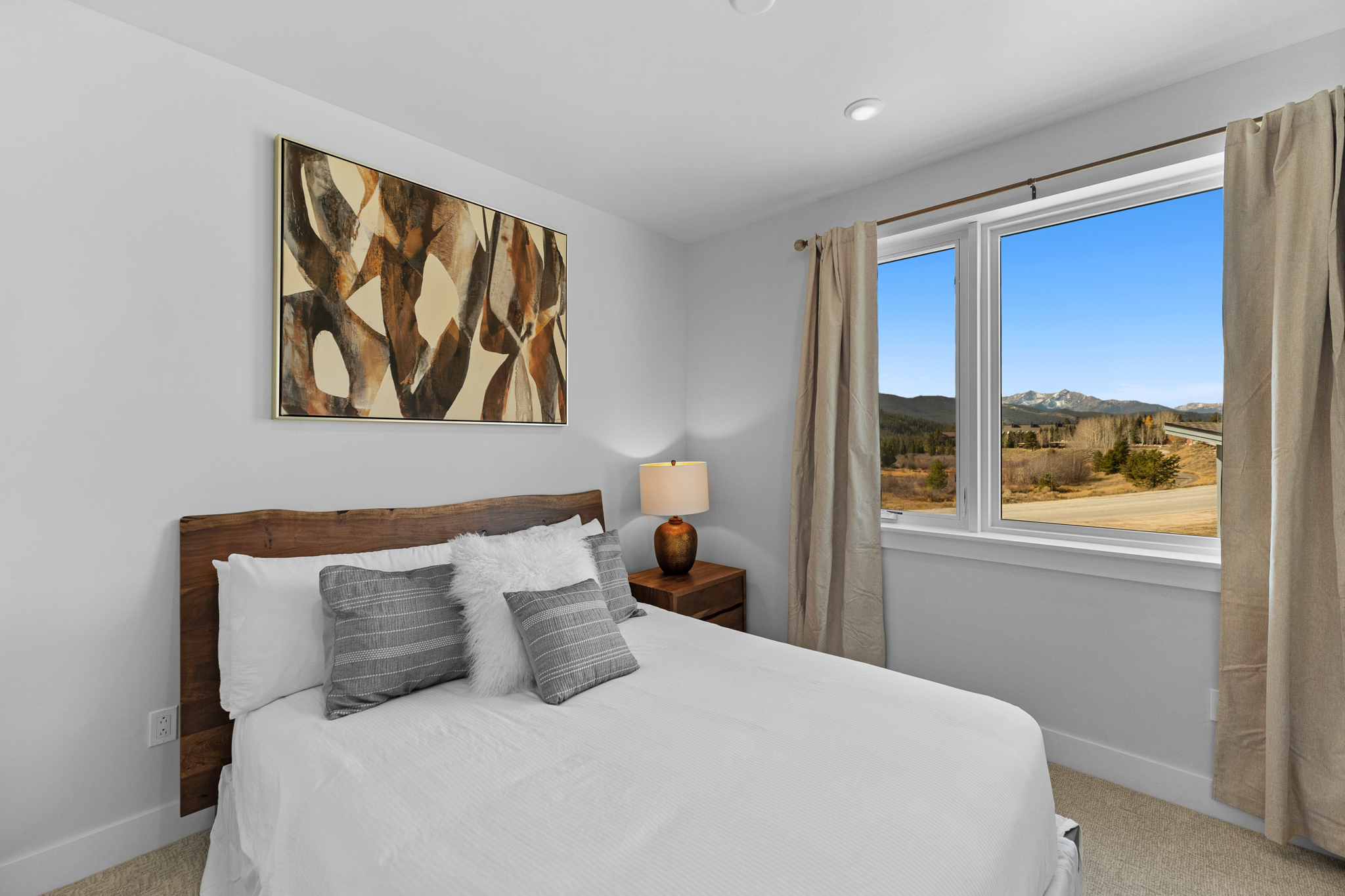 This cozy bedroom features a rustic live-edge wood headboard paired with a crisp white bedspread and decorative throw pillows. A bedside table with a warm-toned lamp sits next to a large window that frames a stunning, expansive view of the mountains and natural landscape. The room is decorated in a neutral, modern style, creating a peaceful and inviting atmosphere.
