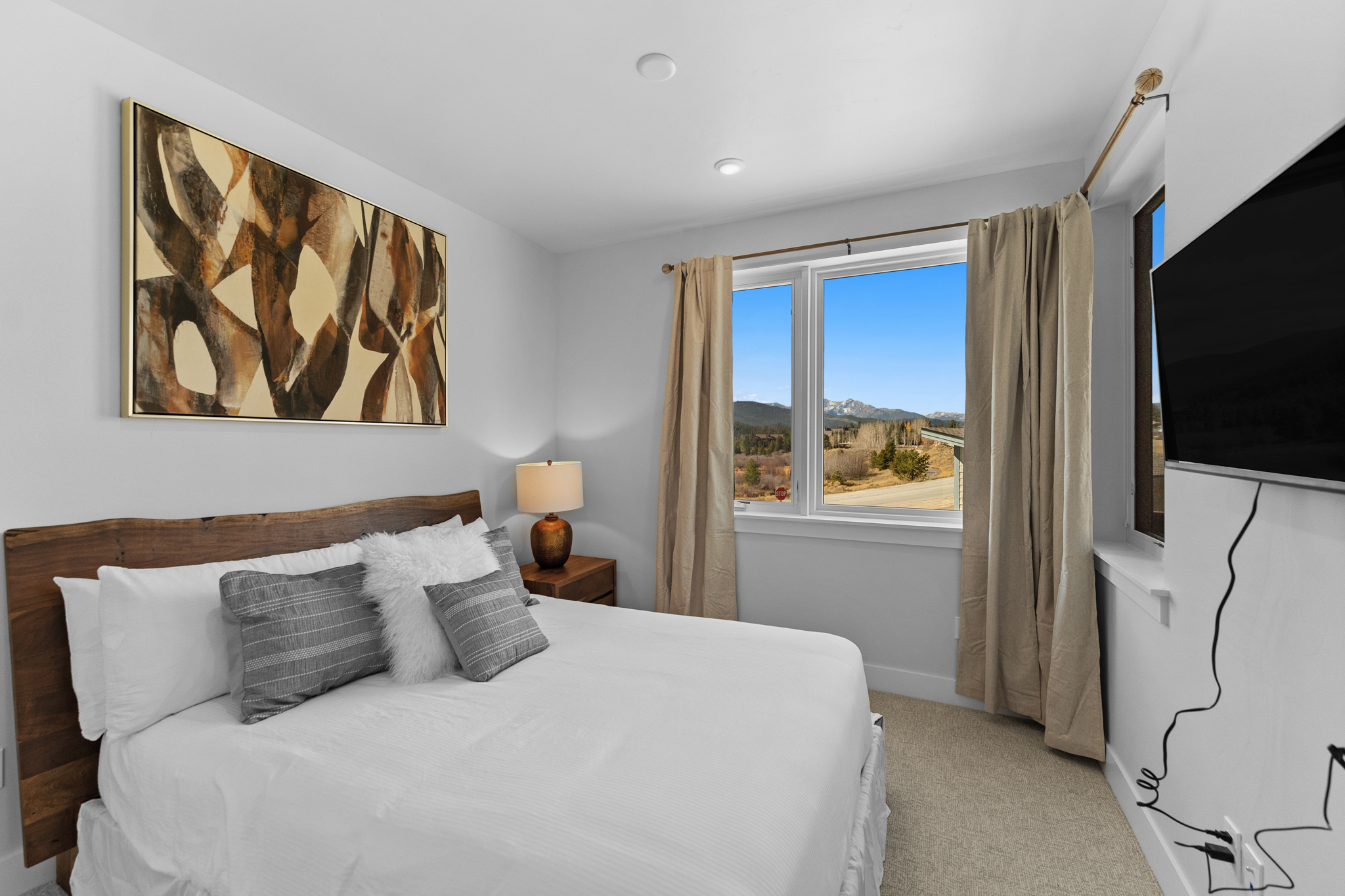 This cozy bedroom features a rustic live-edge wood headboard paired with a crisp white bedspread and decorative gray and white pillows. A large window provides a stunning, clear view of the surrounding mountain landscape, while a wall-mounted television and a bedside lamp add functional comfort. The room is decorated in a neutral, modern style that emphasizes the natural beauty visible outside.