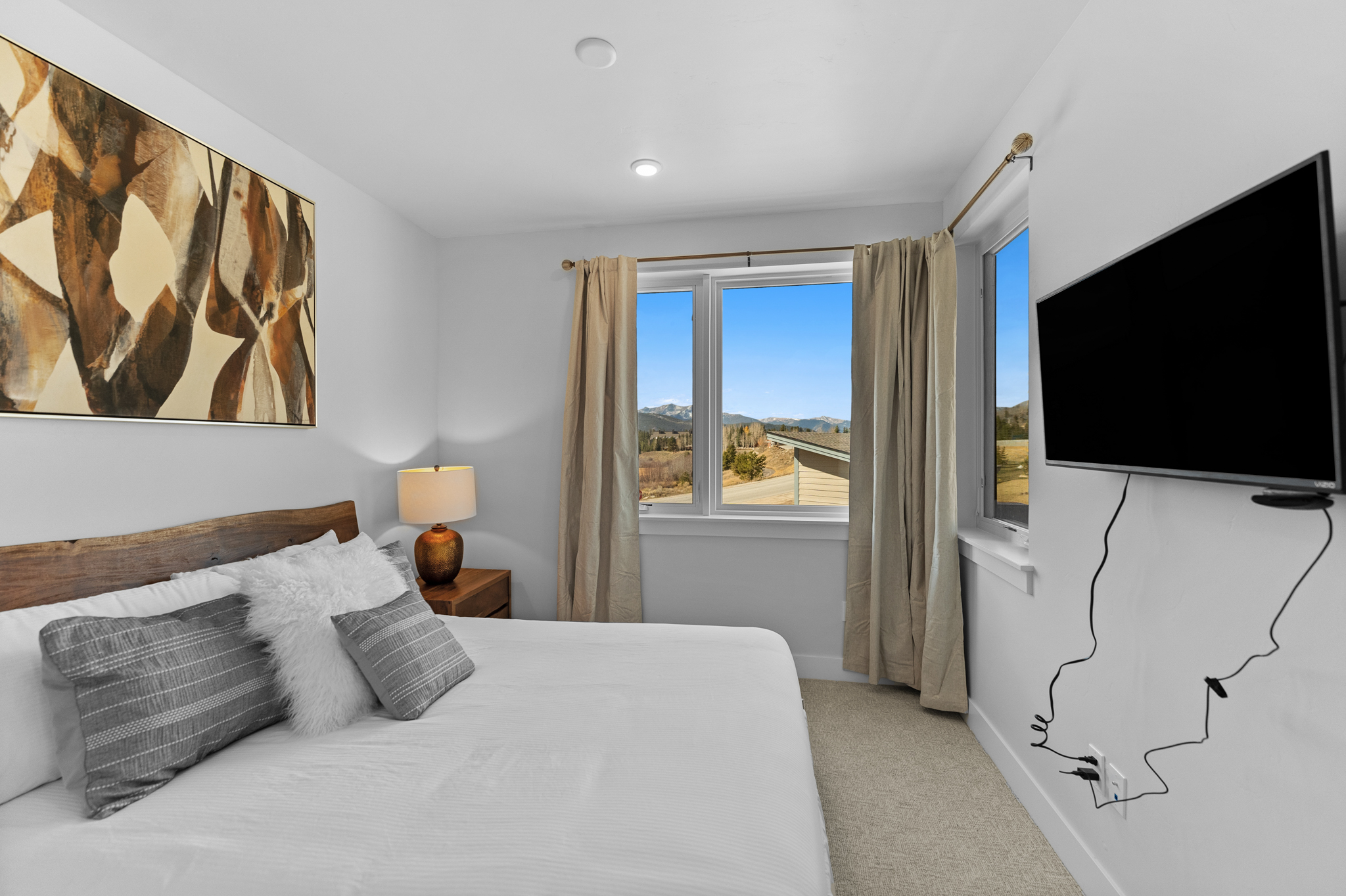 This cozy bedroom features a king-sized bed with a rustic wood headboard, neutral bedding, and decorative pillows. A large window provides a scenic view of the surrounding mountains, while a wall-mounted television and a bedside lamp add functional comfort. The room is decorated in a clean, contemporary style with soft lighting and a neutral color palette.