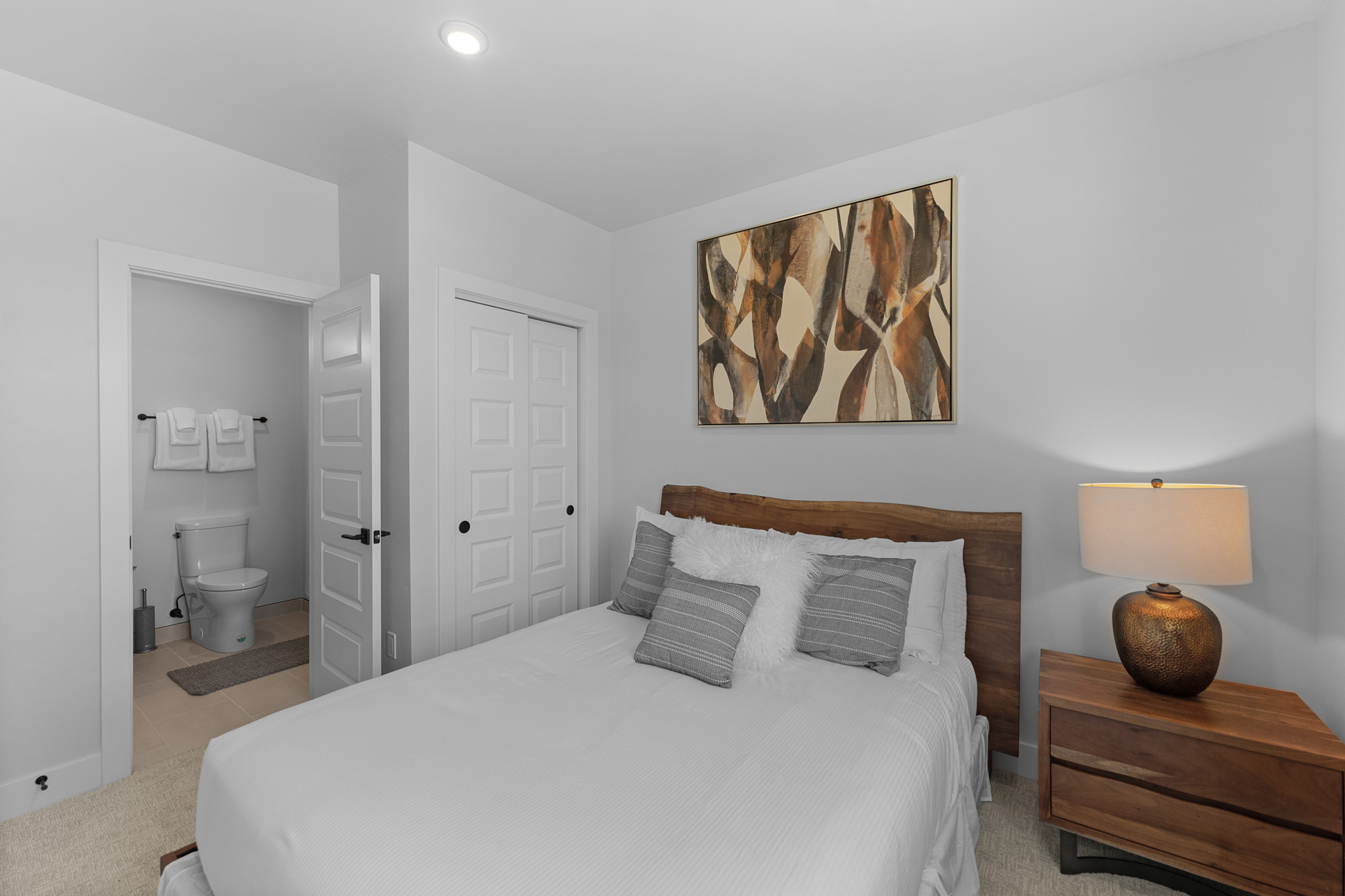 This cozy guest bedroom features a live-edge wooden headboard, a white bedspread with decorative pillows, and a matching wooden nightstand with a warm-toned lamp. An abstract piece of wall art hangs above the bed, while a door leads directly into an en-suite bathroom. The room is painted in a neutral, light gray, creating a clean and inviting atmosphere.