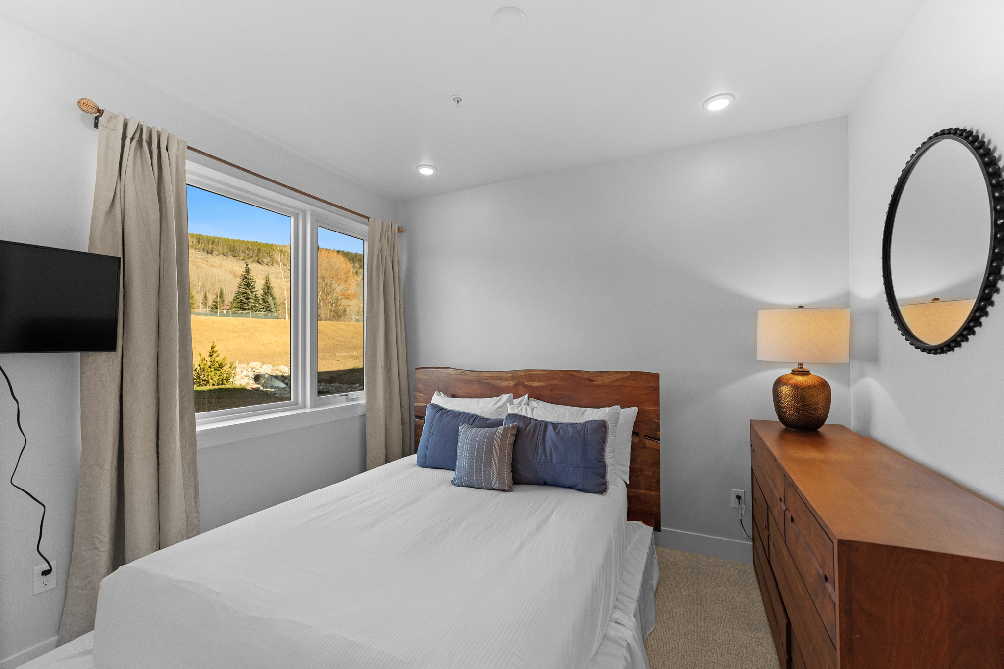 This cozy bedroom features a rustic live-edge wood headboard paired with a white bedspread and blue accent pillows, creating a warm and inviting atmosphere. A wooden dresser with a textured lamp and a circular mirror sits to the right, while a large window provides a scenic view of the surrounding landscape. The room is finished with neutral walls and carpet, offering a clean and comfortable space.