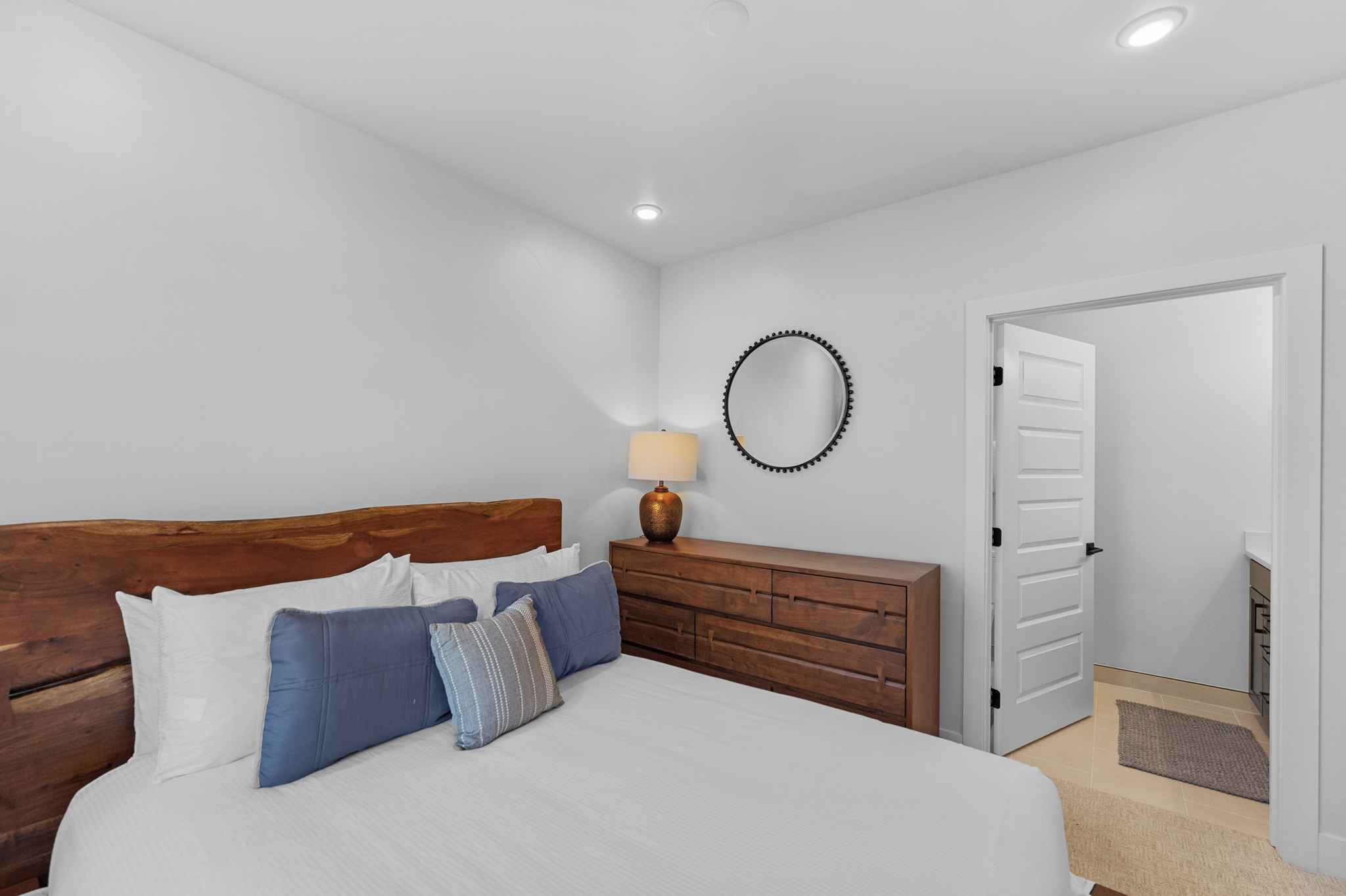 This cozy bedroom features a rustic live-edge wood headboard paired with a matching wooden dresser, creating a warm and inviting atmosphere. A circular mirror hangs above the dresser, while a doorway leads into an adjacent bathroom, suggesting a convenient layout. The room is bright and clean, with a neutral color palette accented by soft blue bedding.