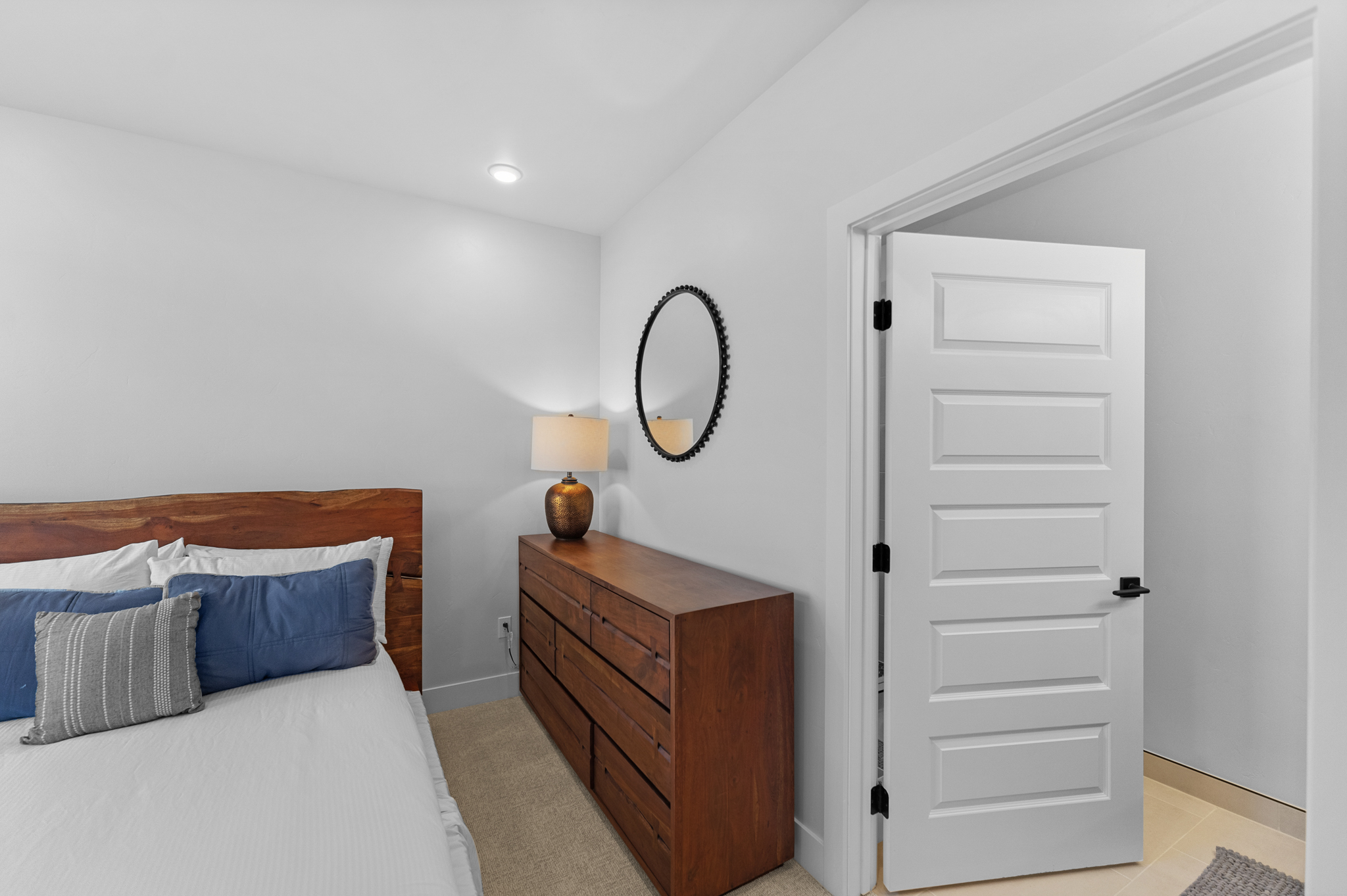 This cozy bedroom features a warm, rustic wooden headboard and a matching dresser, creating a cohesive and inviting atmosphere. The room is accented with a stylish oval mirror and a bedside lamp, while a white door leads into an adjacent space, suggesting a functional layout. The neutral color palette and soft lighting contribute to a calm and comfortable living environment.