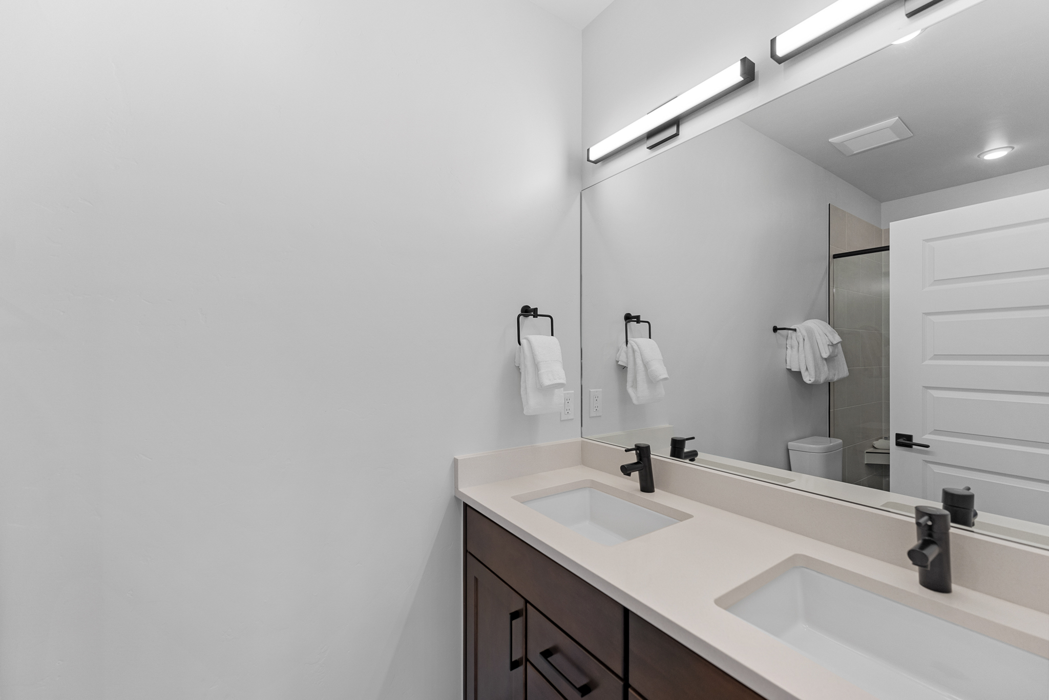 This modern primary bathroom features a double vanity with dark wood cabinetry, a light-colored quartz countertop, and sleek matte black fixtures. A large, frameless mirror spans the wall above the sinks, reflecting the clean, minimalist aesthetic and the adjacent shower area. The space is bright and well-lit, offering a contemporary and functional design suitable for a high-end home.