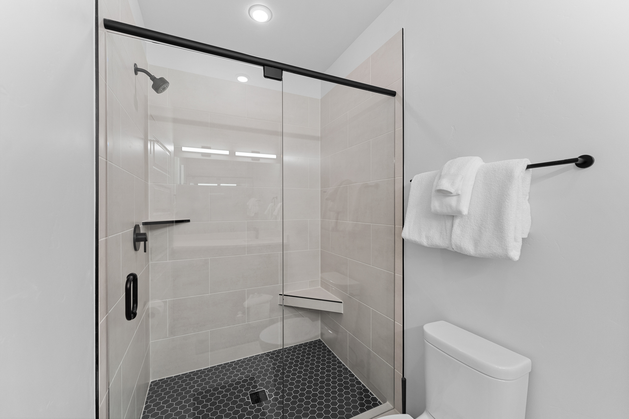 This modern bathroom features a sleek walk-in shower with a black-framed glass enclosure, light-colored subway wall tiles, and a striking black hexagonal tile floor. A built-in corner bench adds functionality to the shower, while a white toilet and a black towel rack are positioned adjacent to the enclosure. The overall aesthetic is clean, contemporary, and high-contrast, emphasizing a minimalist design.