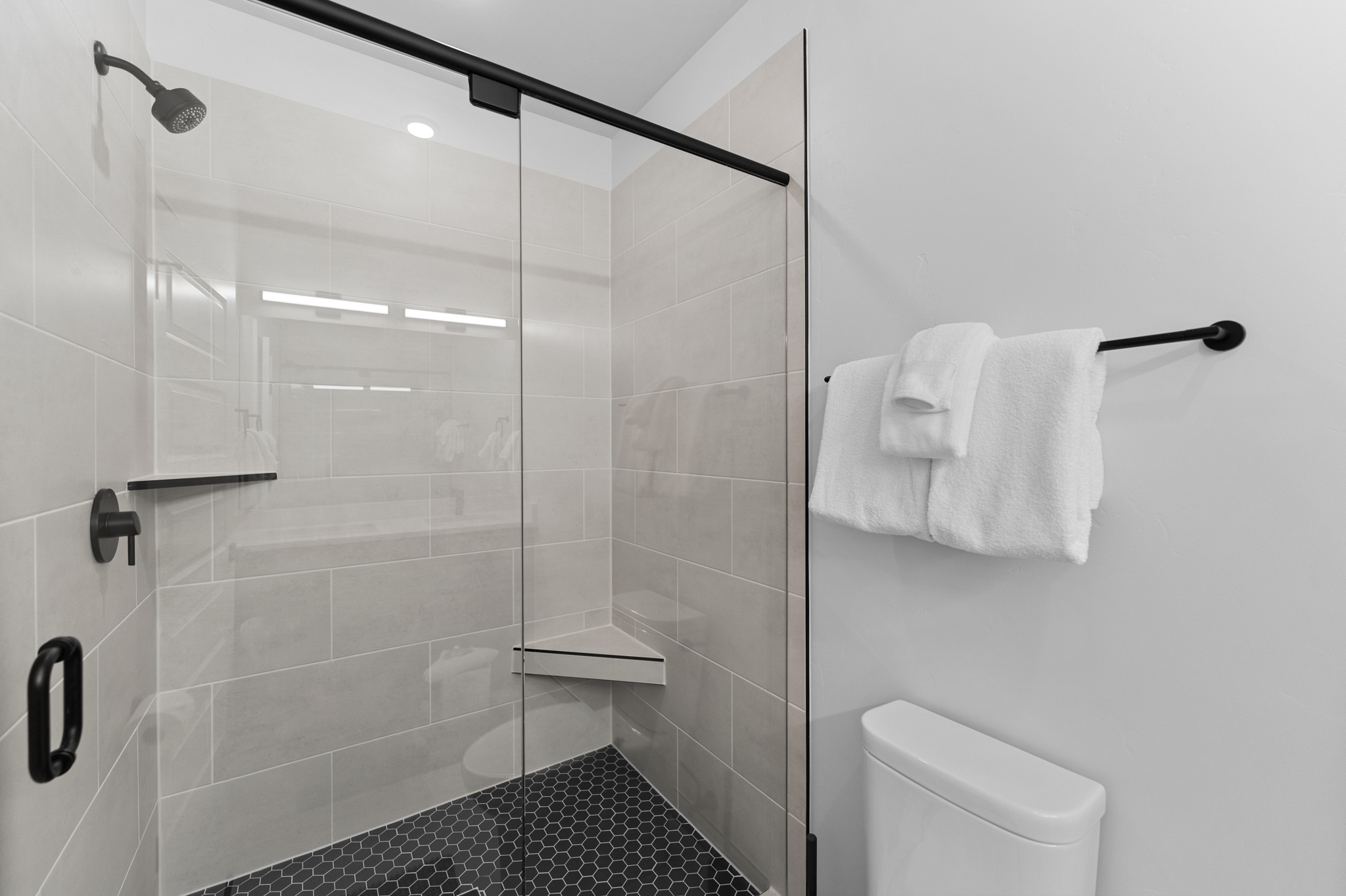 This modern bathroom features a sleek walk-in shower with a glass enclosure, black matte fixtures, and a striking black hexagonal tile floor. The shower includes a built-in corner bench and light-colored subway wall tiles, creating a clean, contemporary aesthetic. A white towel hangs on a black wall-mounted rack next to a minimalist toilet, emphasizing a bright and functional design.