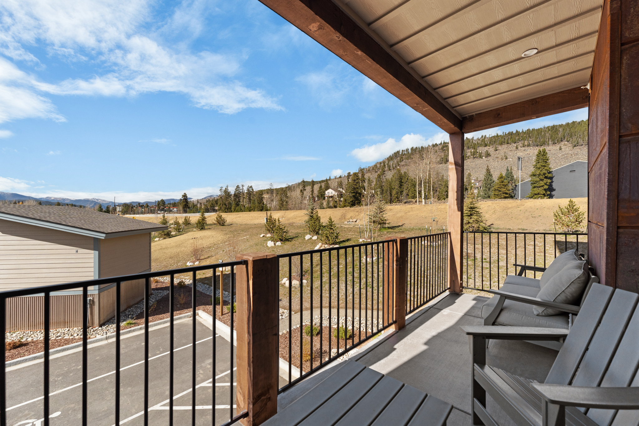This elevated balcony offers a serene, panoramic view of a mountainous landscape and a quiet parking area below. The space is furnished with comfortable outdoor seating, featuring a modern, clean design with wooden support beams and a black metal railing. The perspective captures the inviting atmosphere of a mountain retreat, emphasizing the blend of indoor comfort and expansive natural scenery.