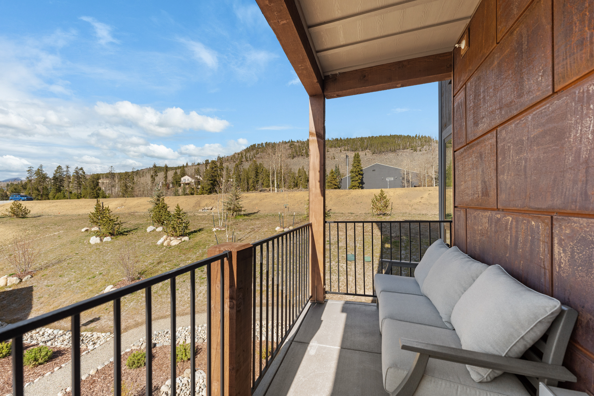 This balcony offers a serene, elevated view of a rolling landscape with distant mountains and a clear blue sky. The space is furnished with a comfortable, light-gray cushioned sofa, set against a rustic, weathered metal-paneled wall and a sturdy wooden support beam. The black metal railing provides a clean, modern contrast to the natural, rugged scenery beyond.
