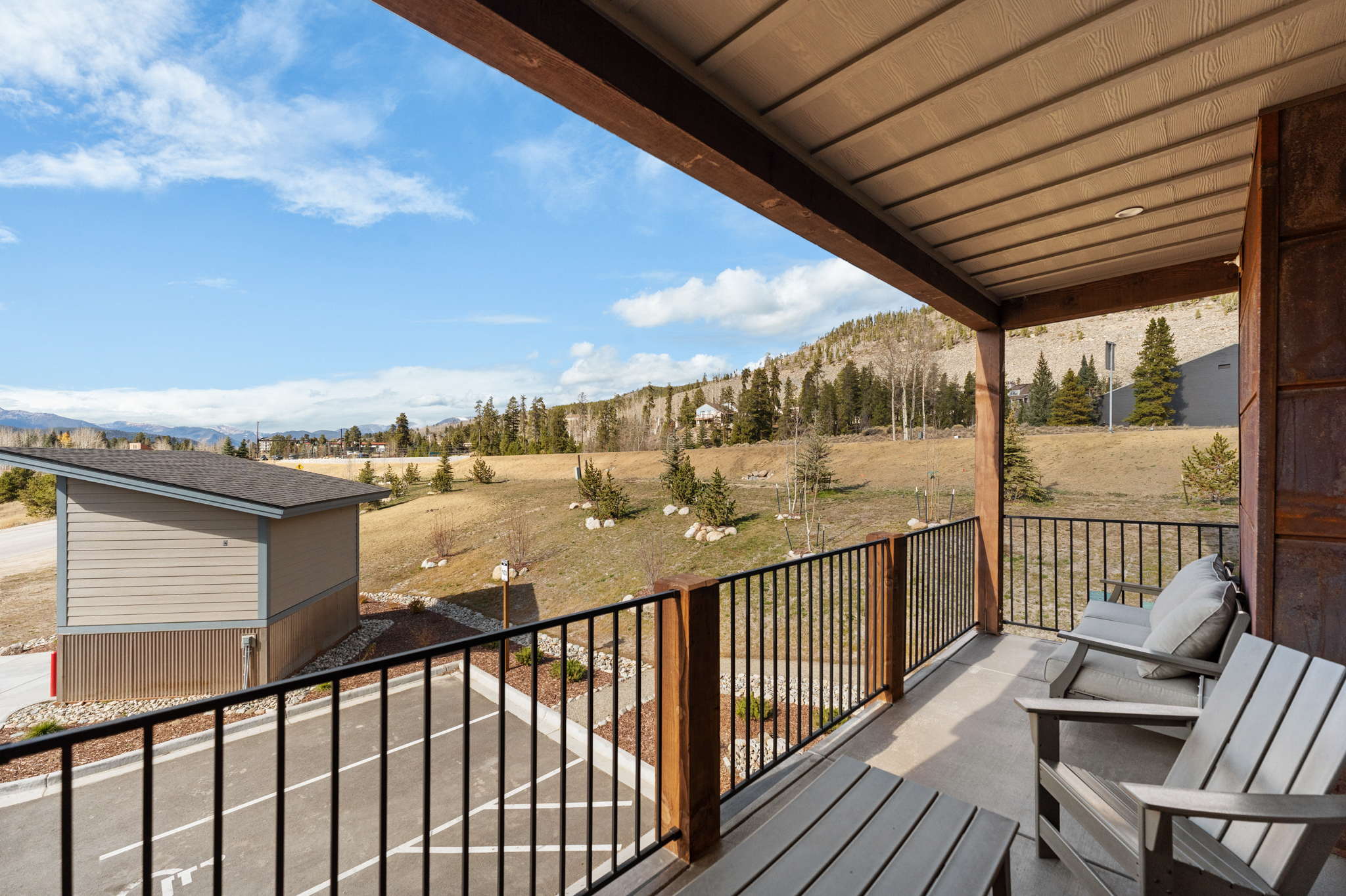 This elevated balcony offers a serene, panoramic view of a mountainous landscape and a quiet parking area below. The space is furnished with comfortable outdoor seating, including a wooden chair and a cushioned sofa, perfect for relaxing and taking in the scenery. The architectural design features a rustic wooden support beam and a clean, modern metal railing, creating a welcoming and peaceful outdoor retreat.