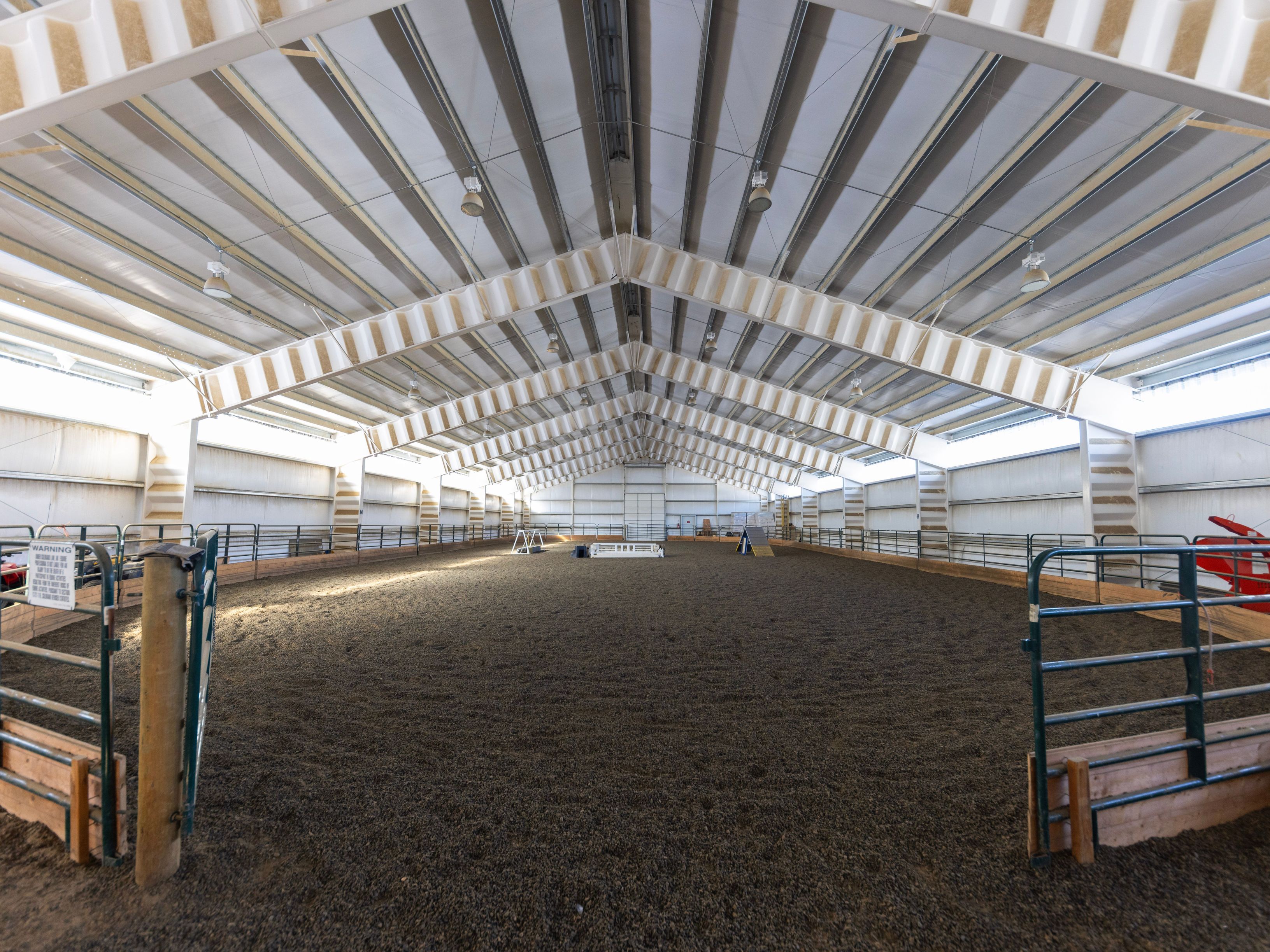 This image showcases a spacious indoor equestrian riding arena with a large, open dirt floor and high, vaulted ceilings supported by industrial steel trusses. The perimeter is enclosed by metal fencing and wooden kickboards, creating a functional and well-maintained training environment. The perspective is a wide-angle shot from the entrance, emphasizing the vast scale and professional utility of the facility.